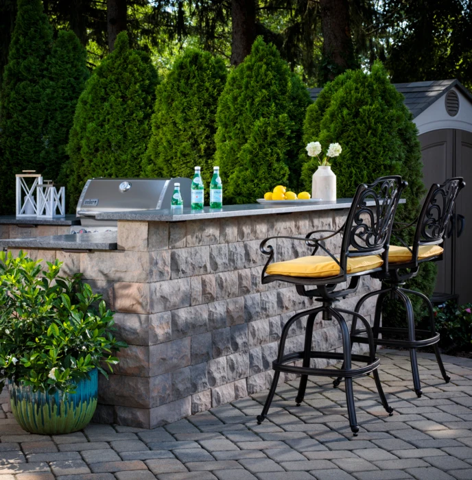 Stone outdoor bar with a built-in grill, stone countertop and two bar stools on a paver patio