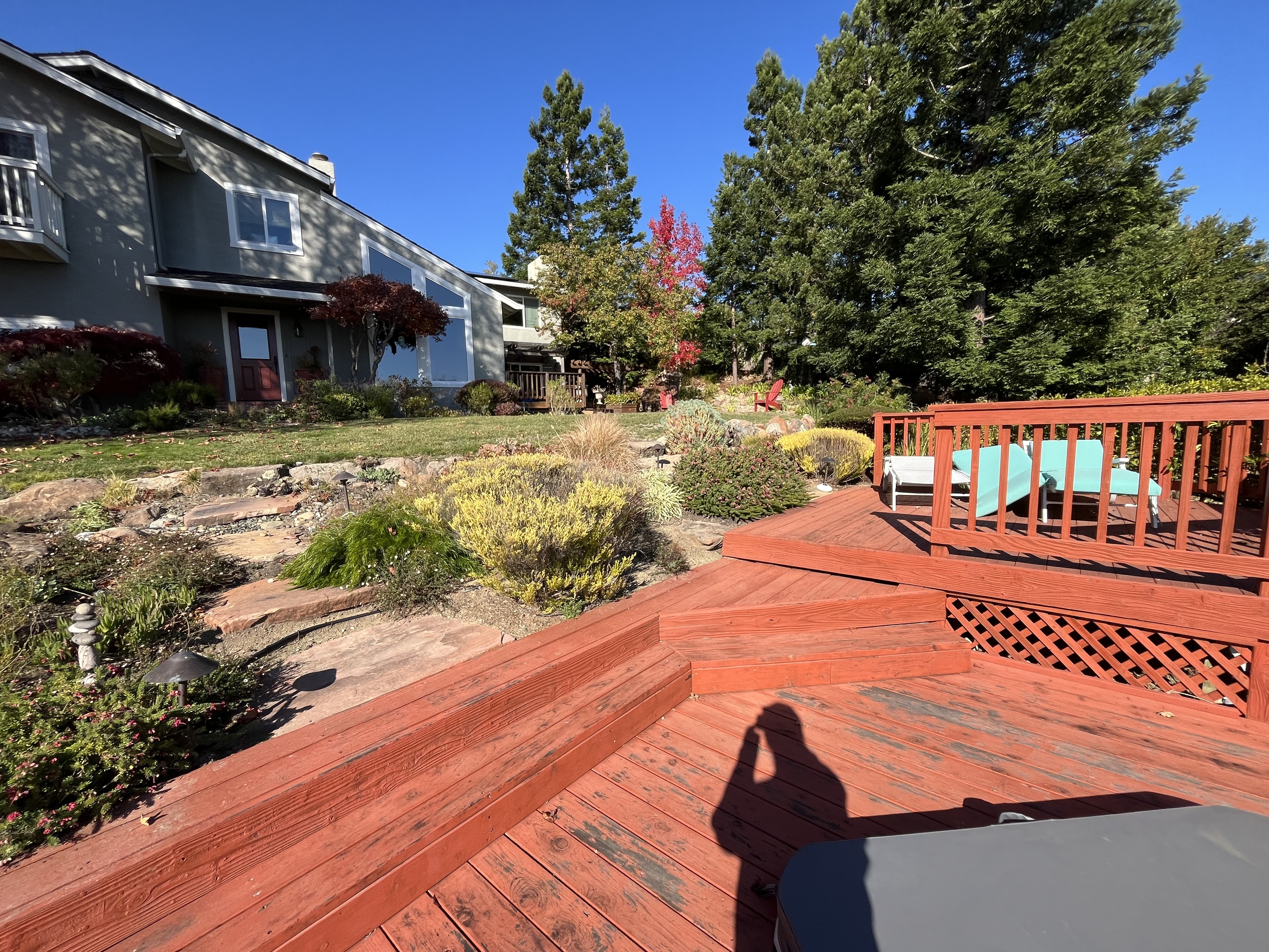 Residential backyard shows a red-stained wooden deck with railing and lattice skirt overlooking terraced rock landscaping...