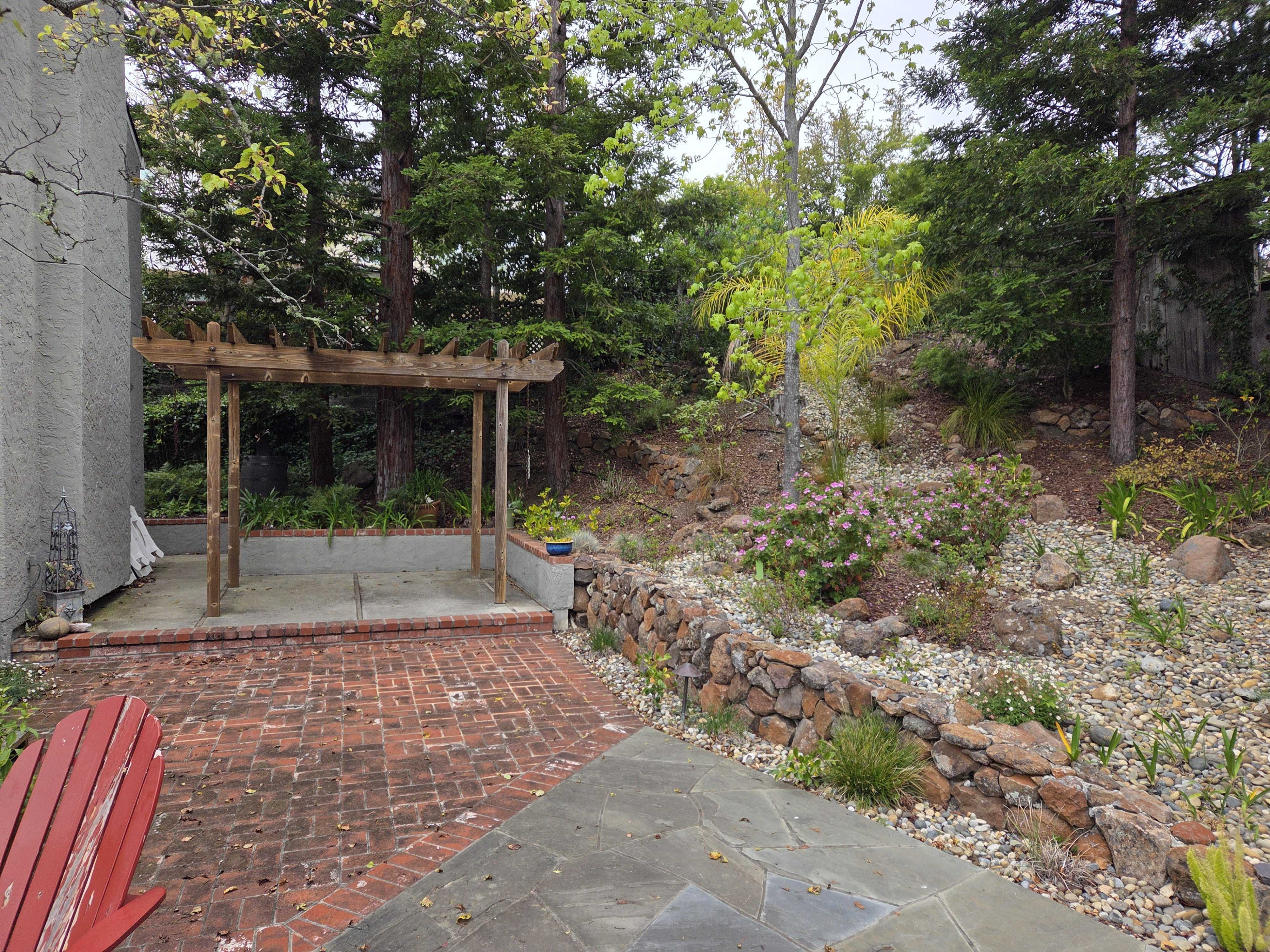 Residential backyard showing a red brick paver patio leading to a small concrete pergola pad, with a natural stacked...