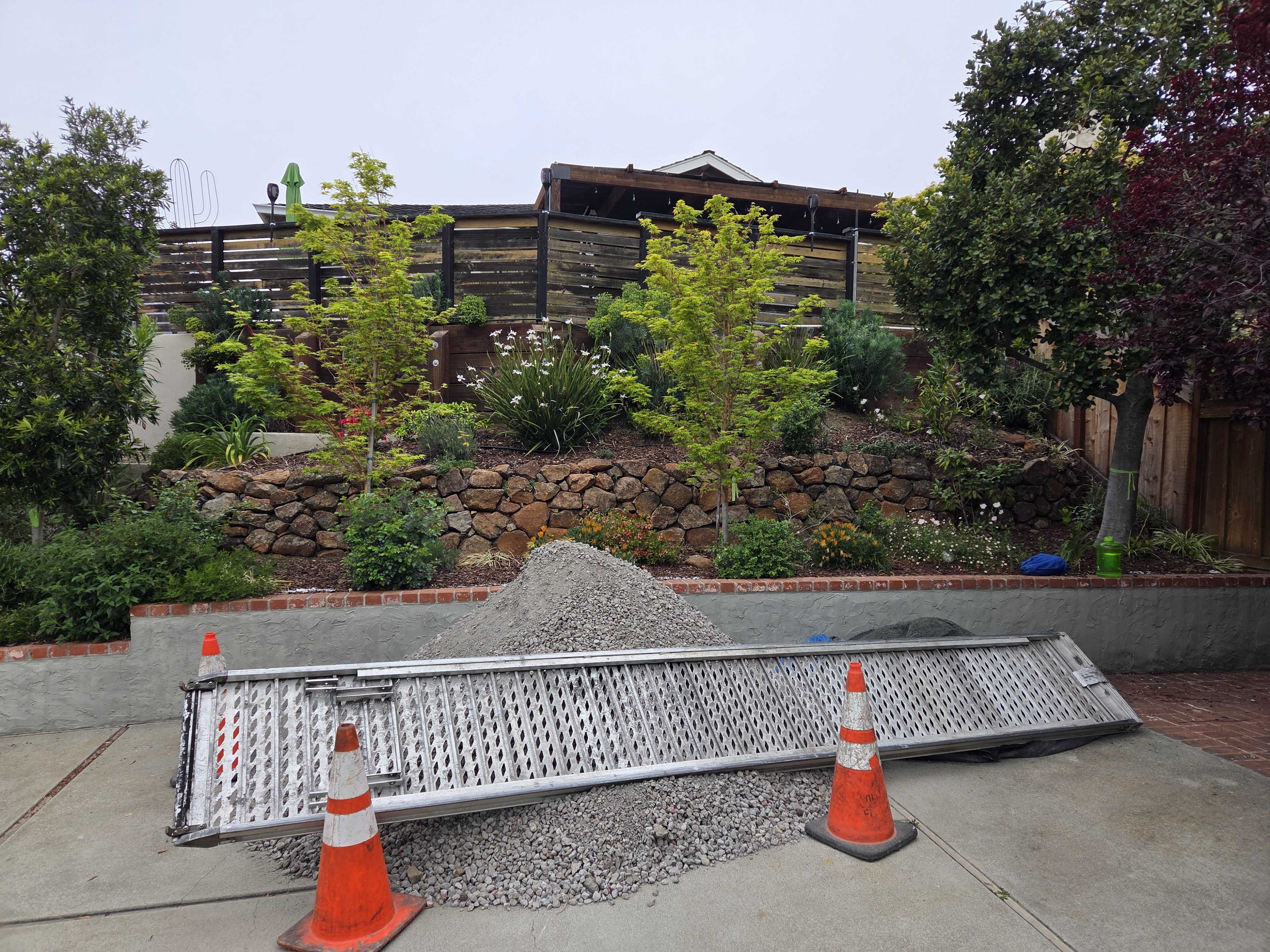 Backyard construction scene showing a piled mound of crushed gravel staged on a concrete driveway/patio area with two...