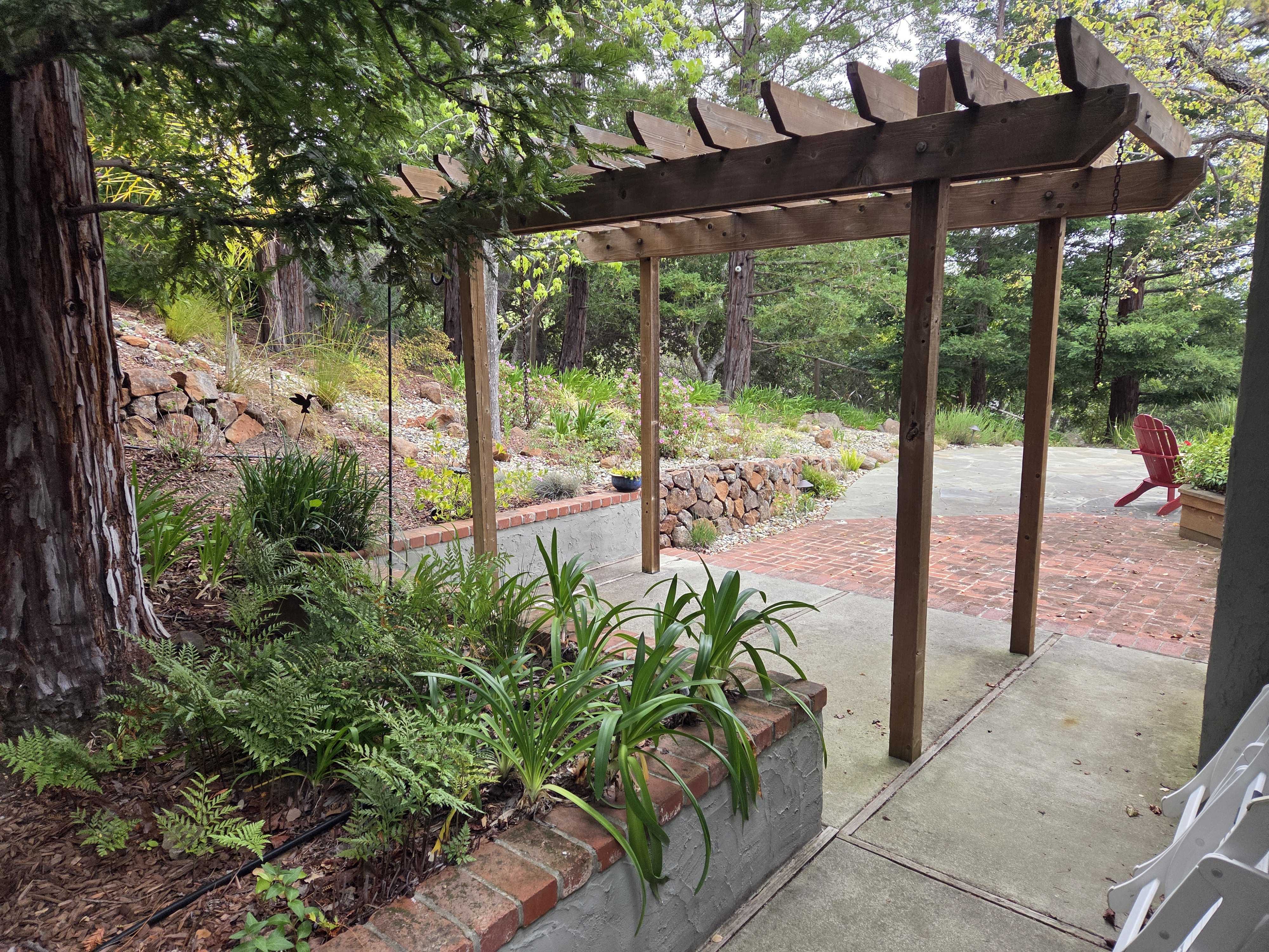 Backyard view showing a wooden pergola over a concrete walkway that leads to a brick paver patio. A segmental stone...