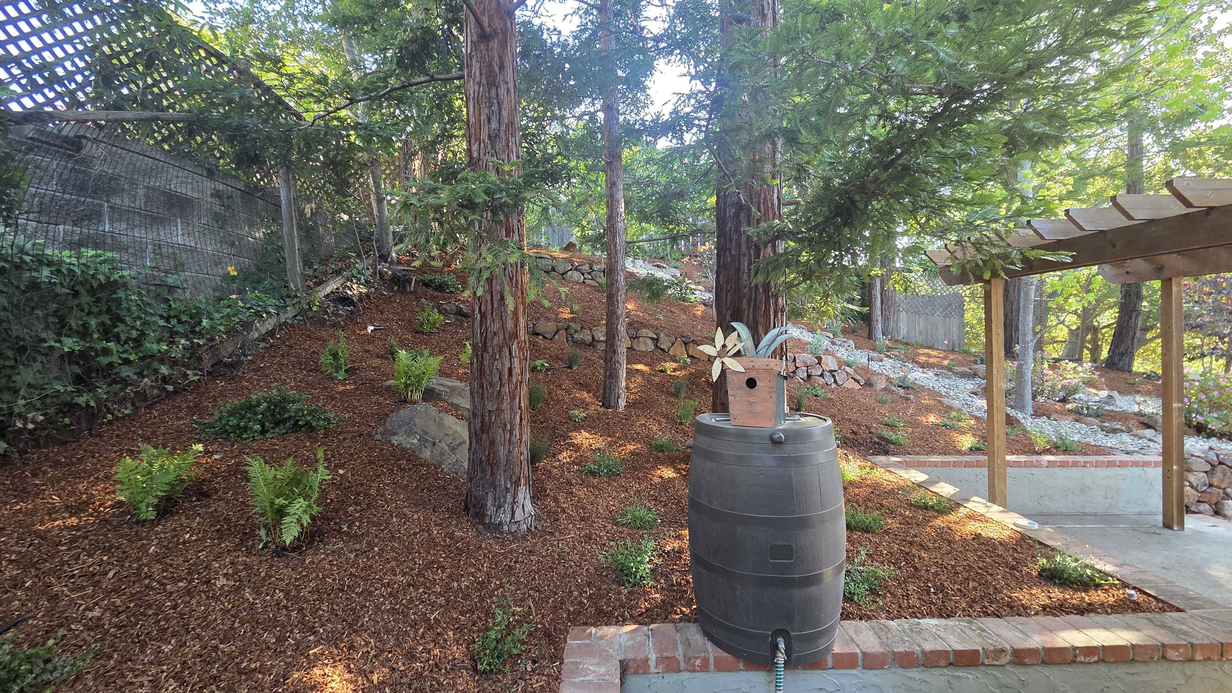 Backyard slope finished with mulch and newly planted shrubs, anchored by a dry-stacked stone retaining wall at the top of...