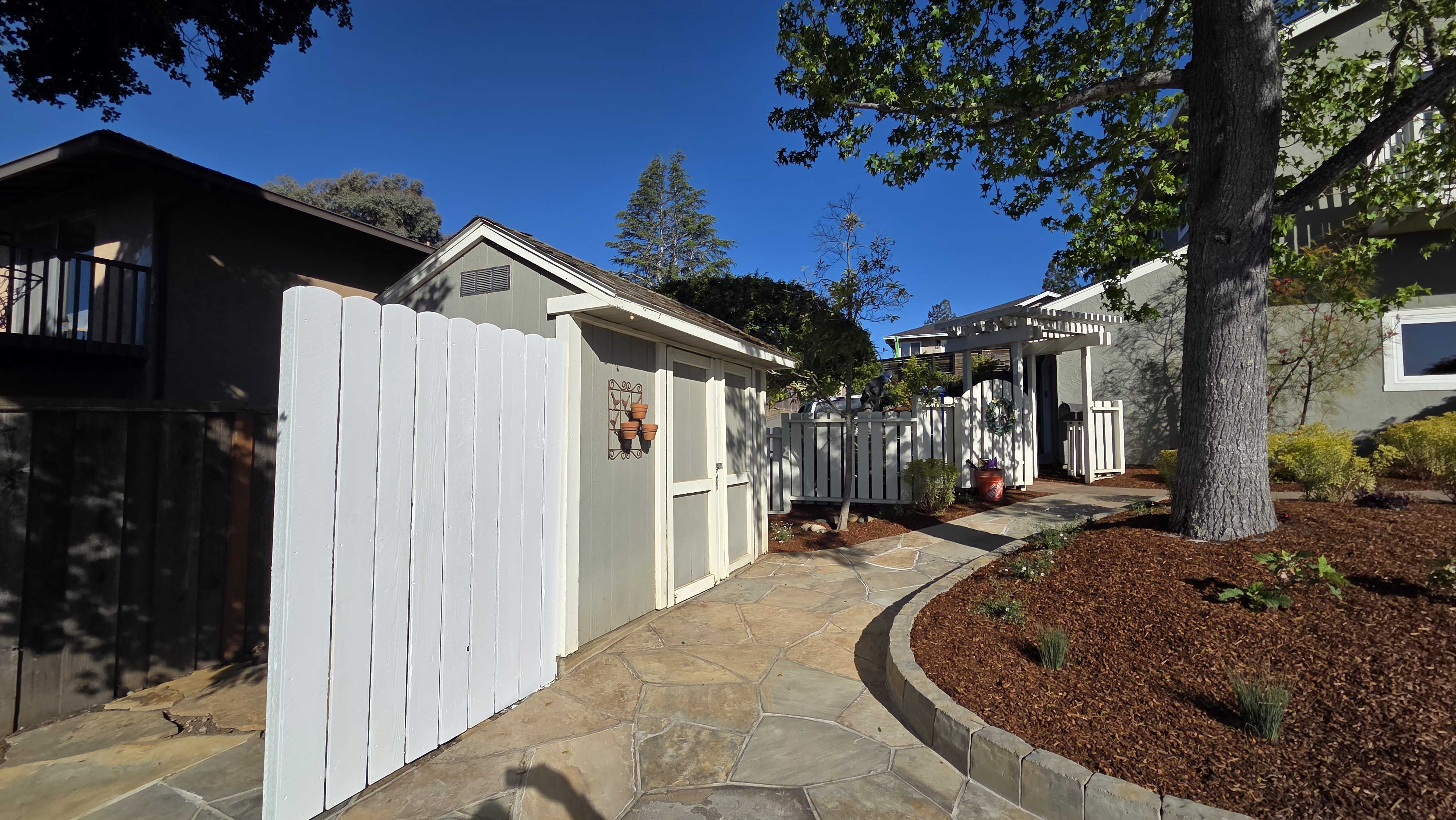 Flagstone paver walkway with a curved concrete/segmental border leading past a small shed and white fence into a...