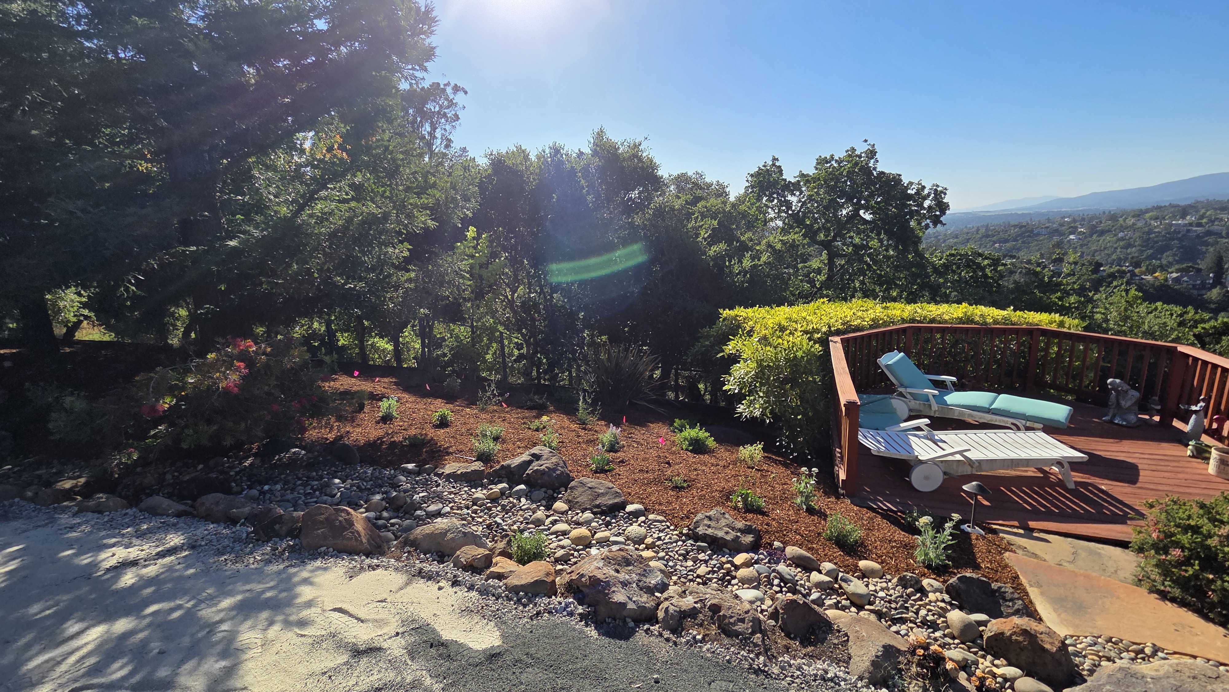 Finished residential backyard showing a wood deck with lounge chairs adjacent to a paved patio area and a landscaped...