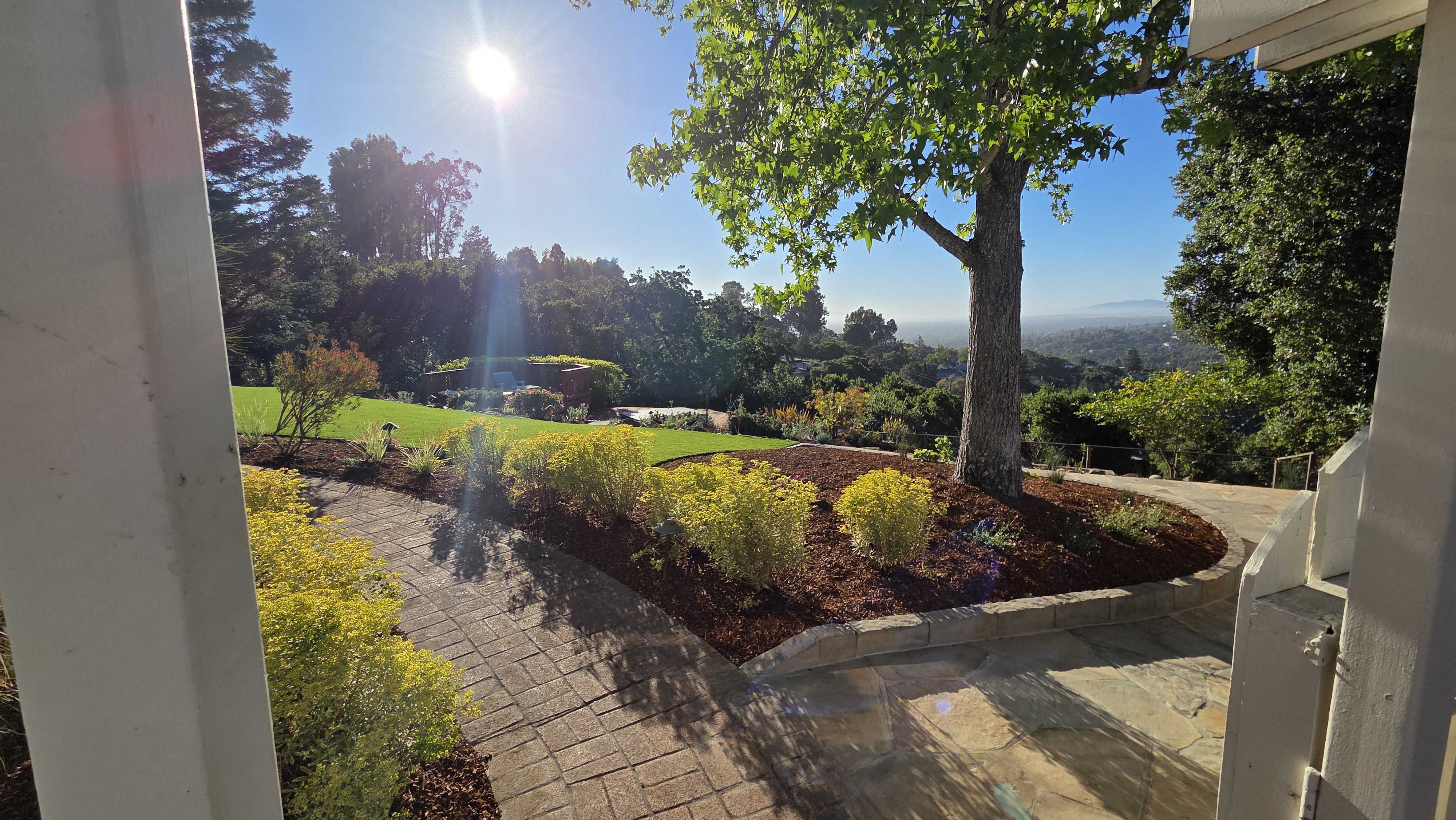 Backyard site photo showing a curved paver walkway and adjoining stone patio opening onto a uniform artificial turf lawn....