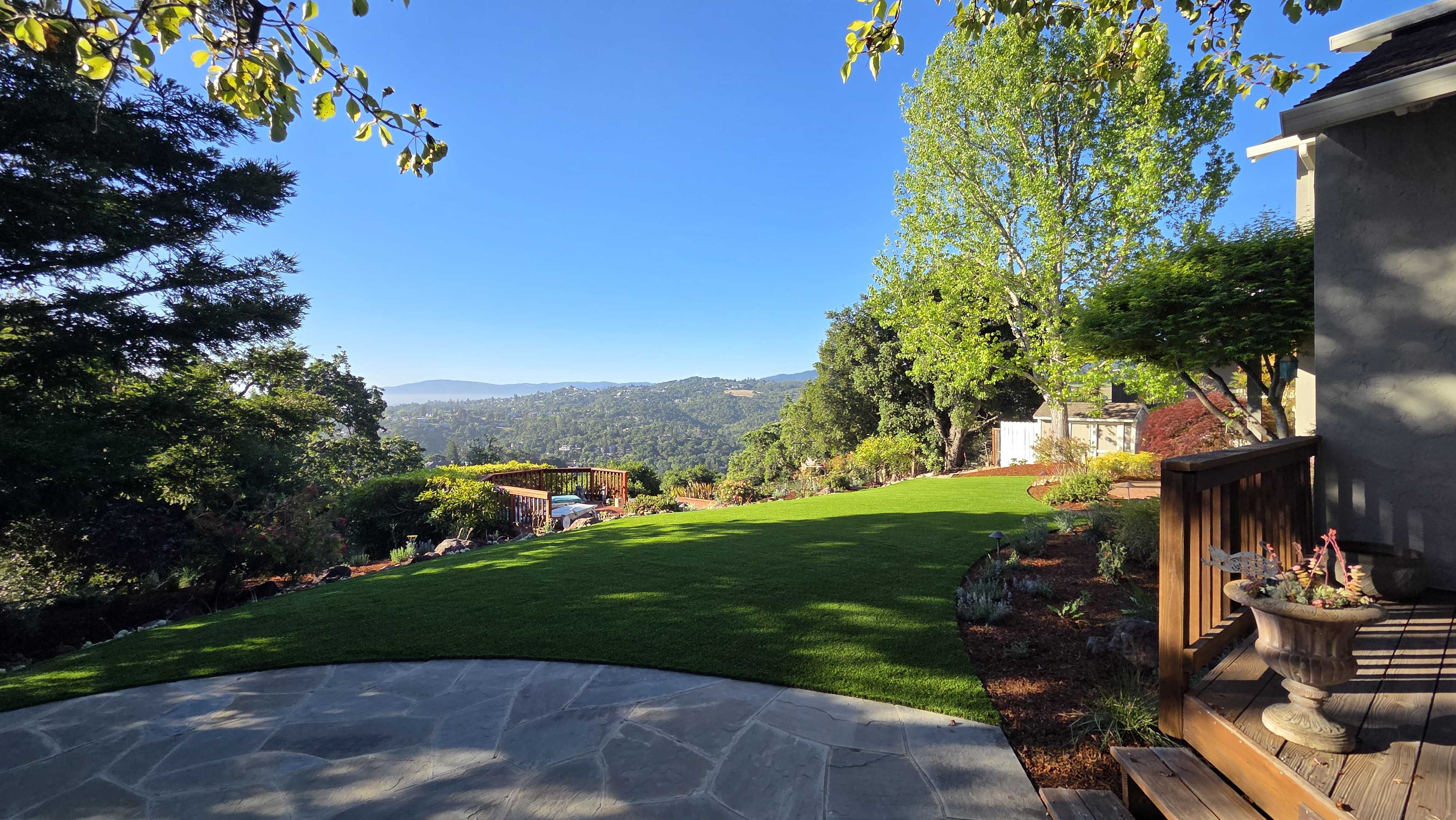 Residential backyard with a curved stone paver patio in the foreground and a newly installed artificial turf lawn...