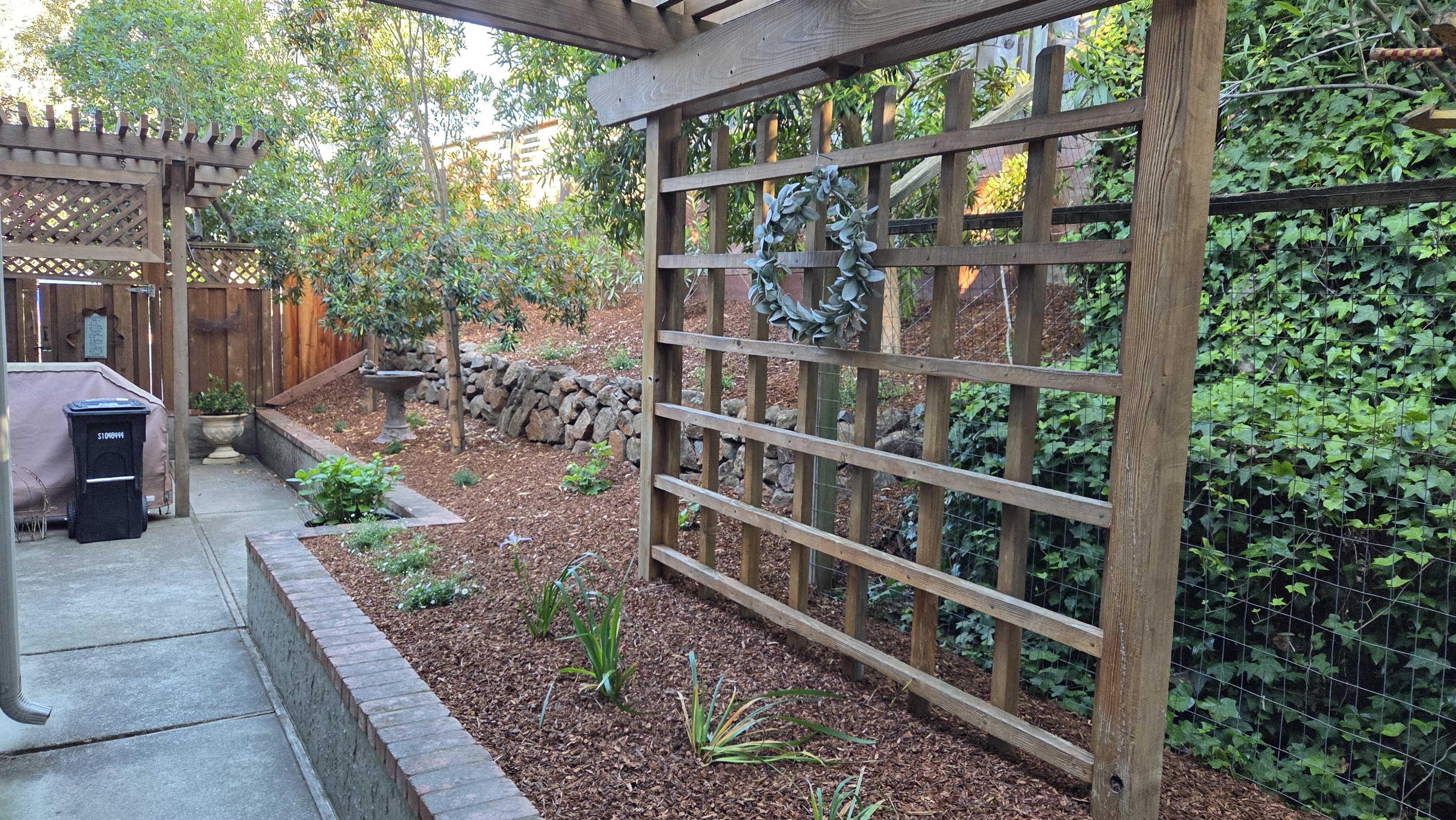Residential side-yard showing a concrete walkway flanking a raised planting bed capped with brick coping and filled with...