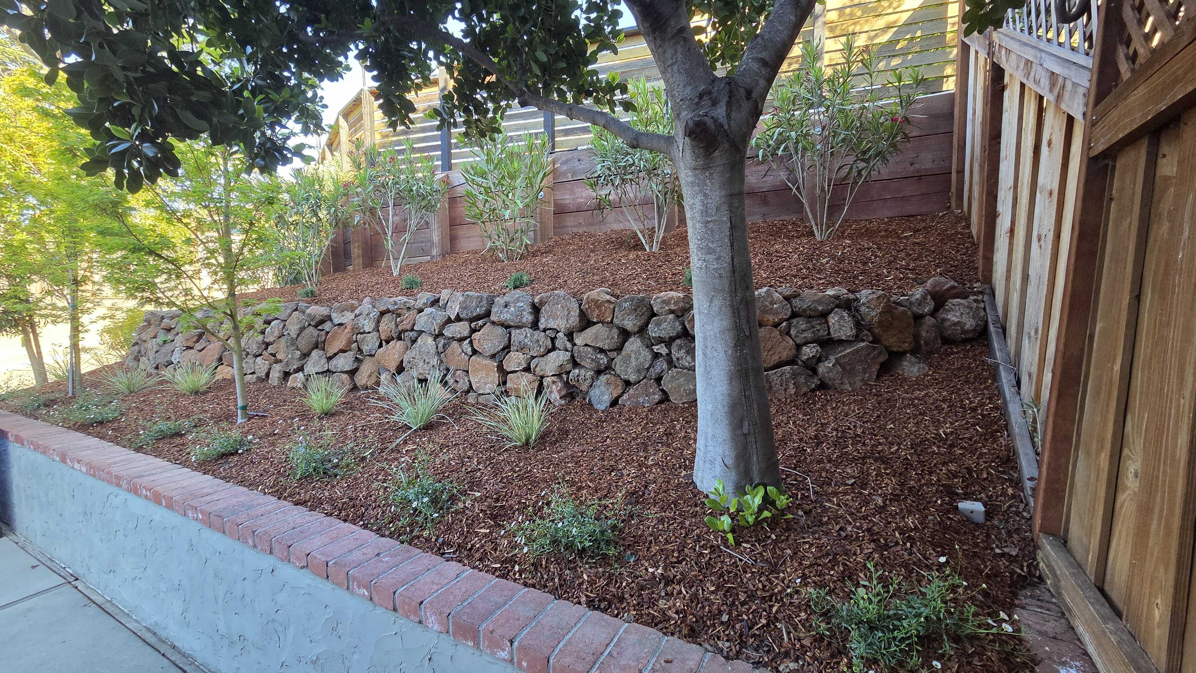 Residential backyard showing a completed slope with a dry-stacked stone retaining wall and a lower concrete knee wall...
