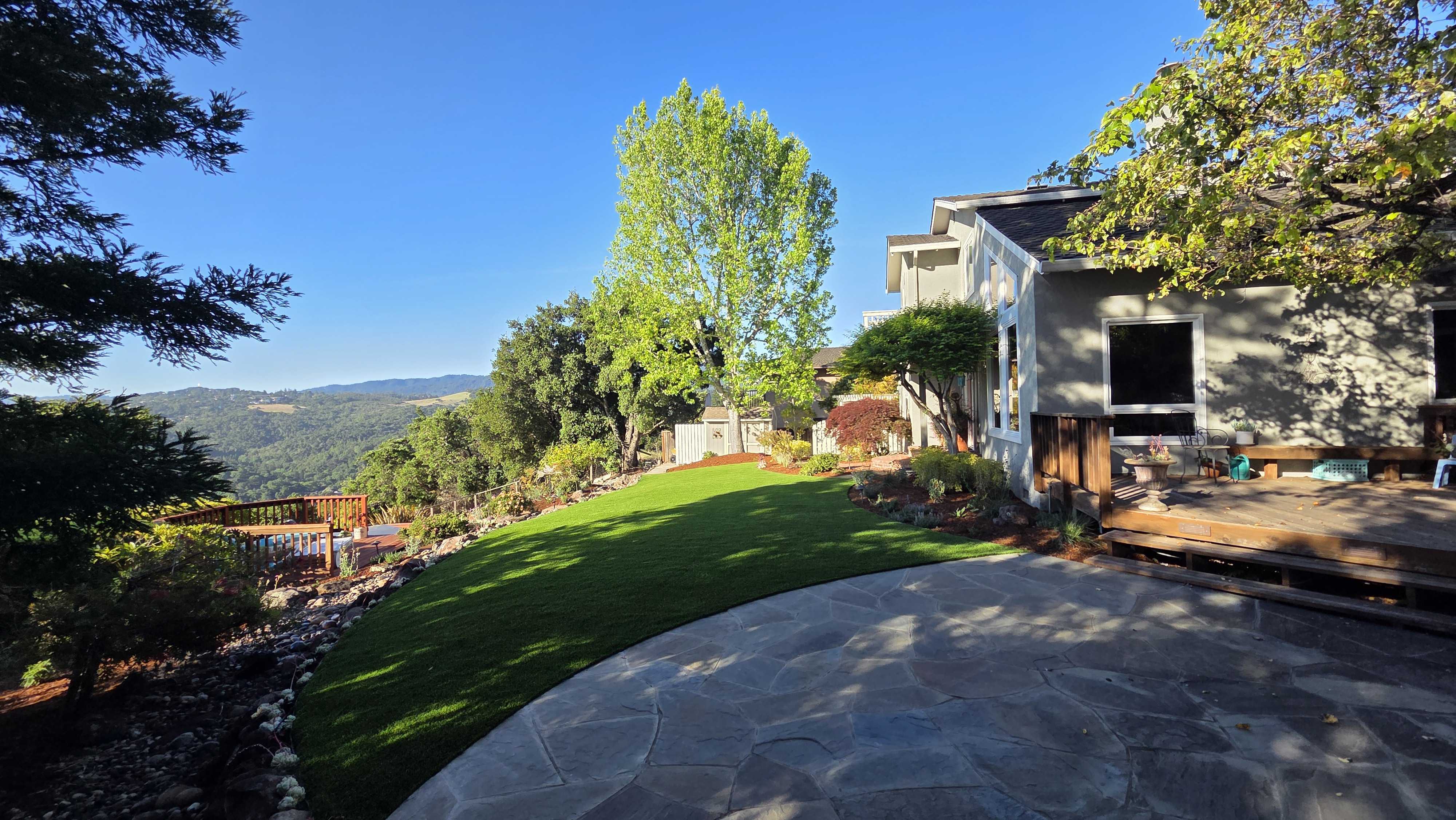 Residential backyard showing a completed outdoor living area with newly installed artificial turf flowing into a natural...