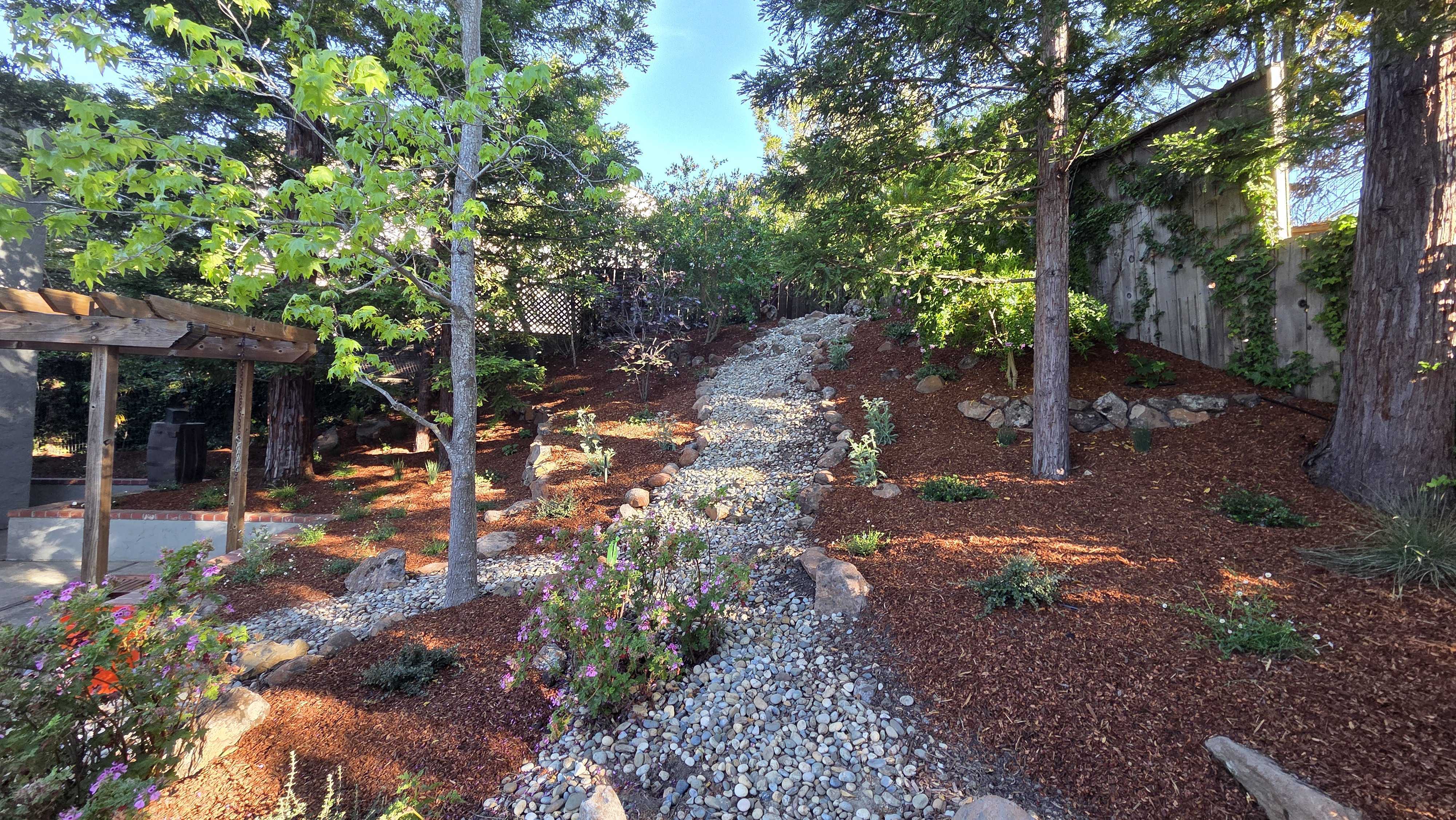 Finished residential backyard landscaping showing a sloped mulch bed with a decorative dry creekbed of river rock winding...