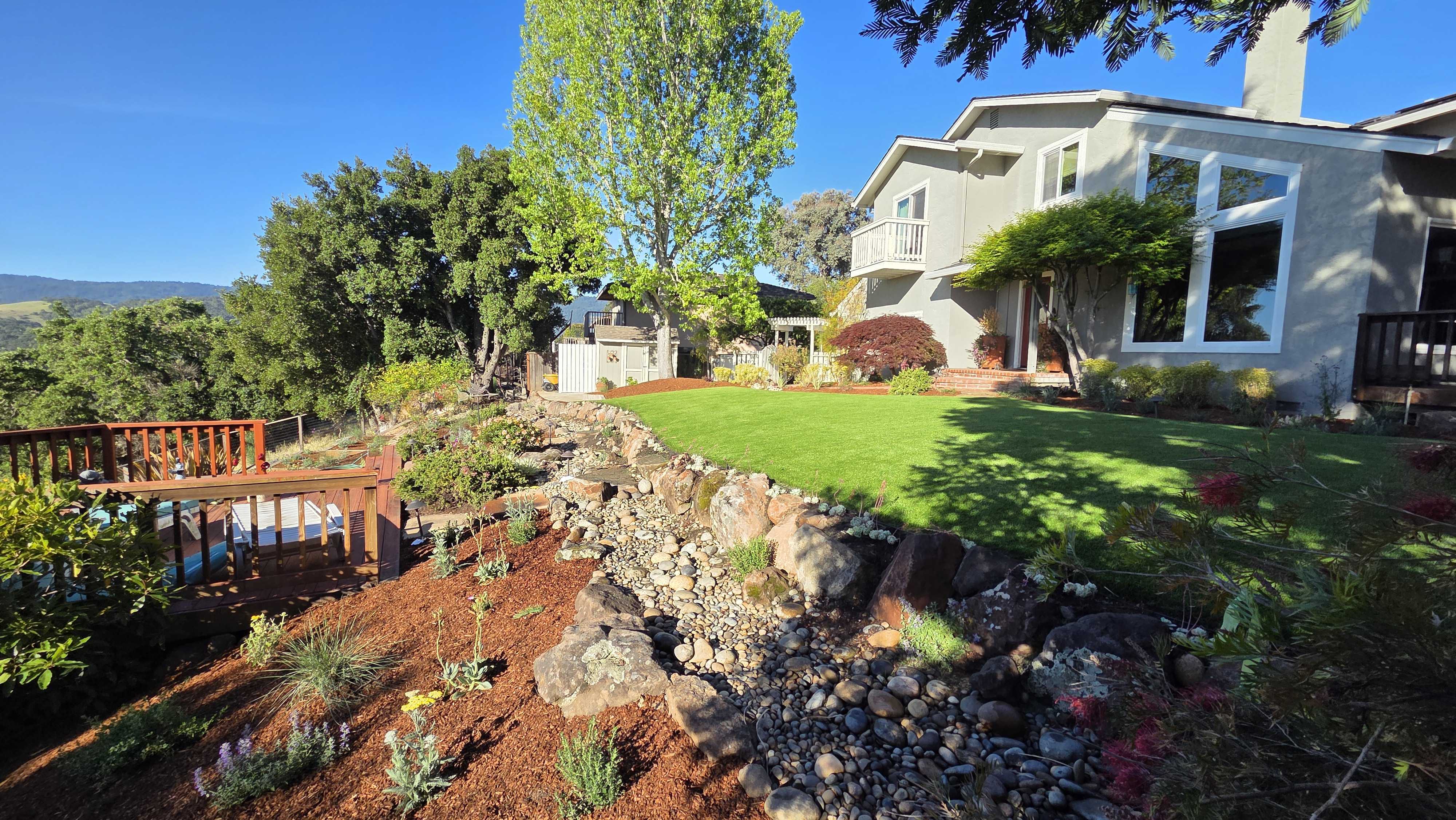 Residential backyard showing a finished artificial turf lawn above a rock retaining wall with a river-rock drainage swale...