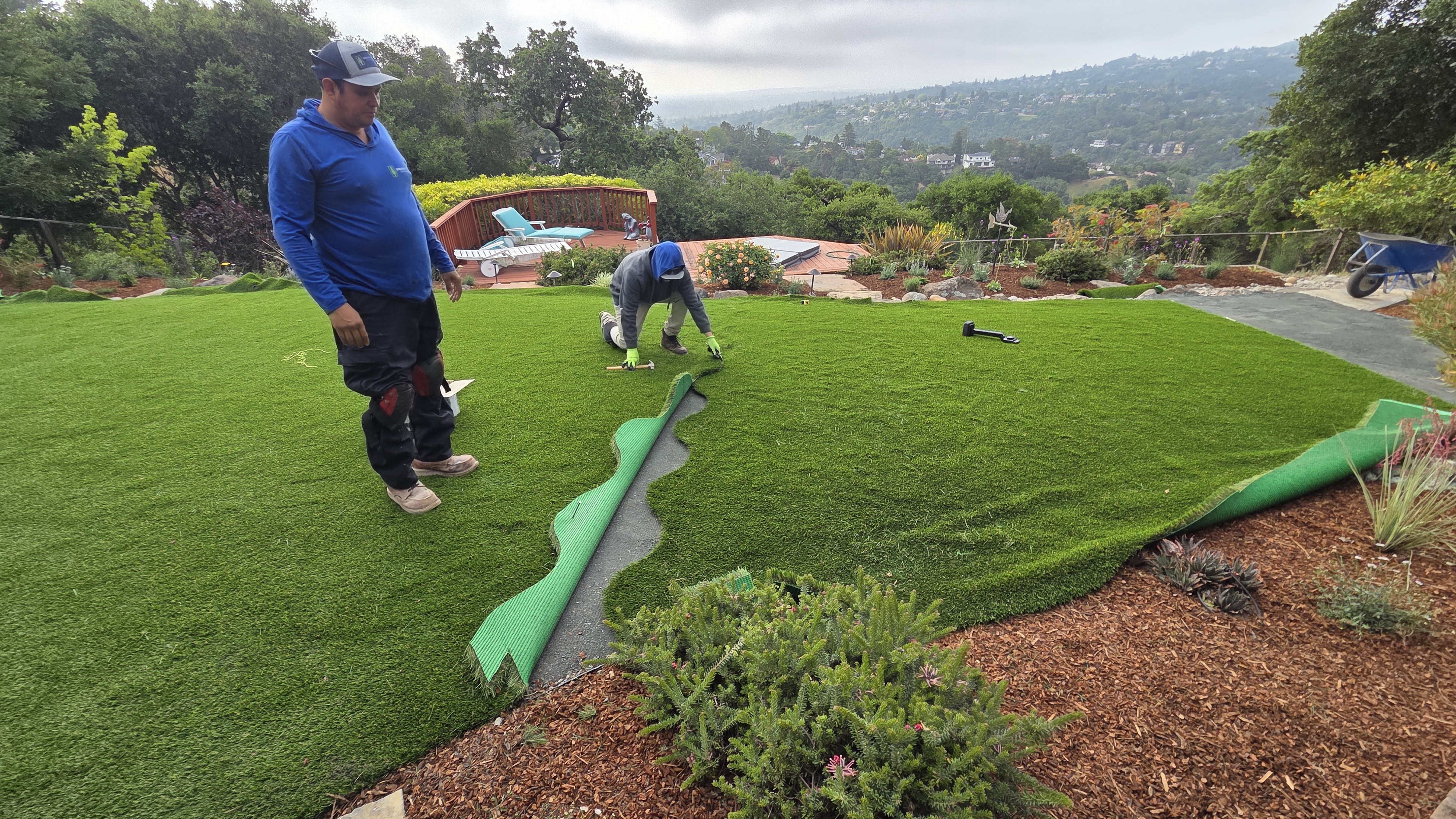 Two crew members are installing synthetic turf on a residential backyard slope; a seam and turf underlayment are exposed...
