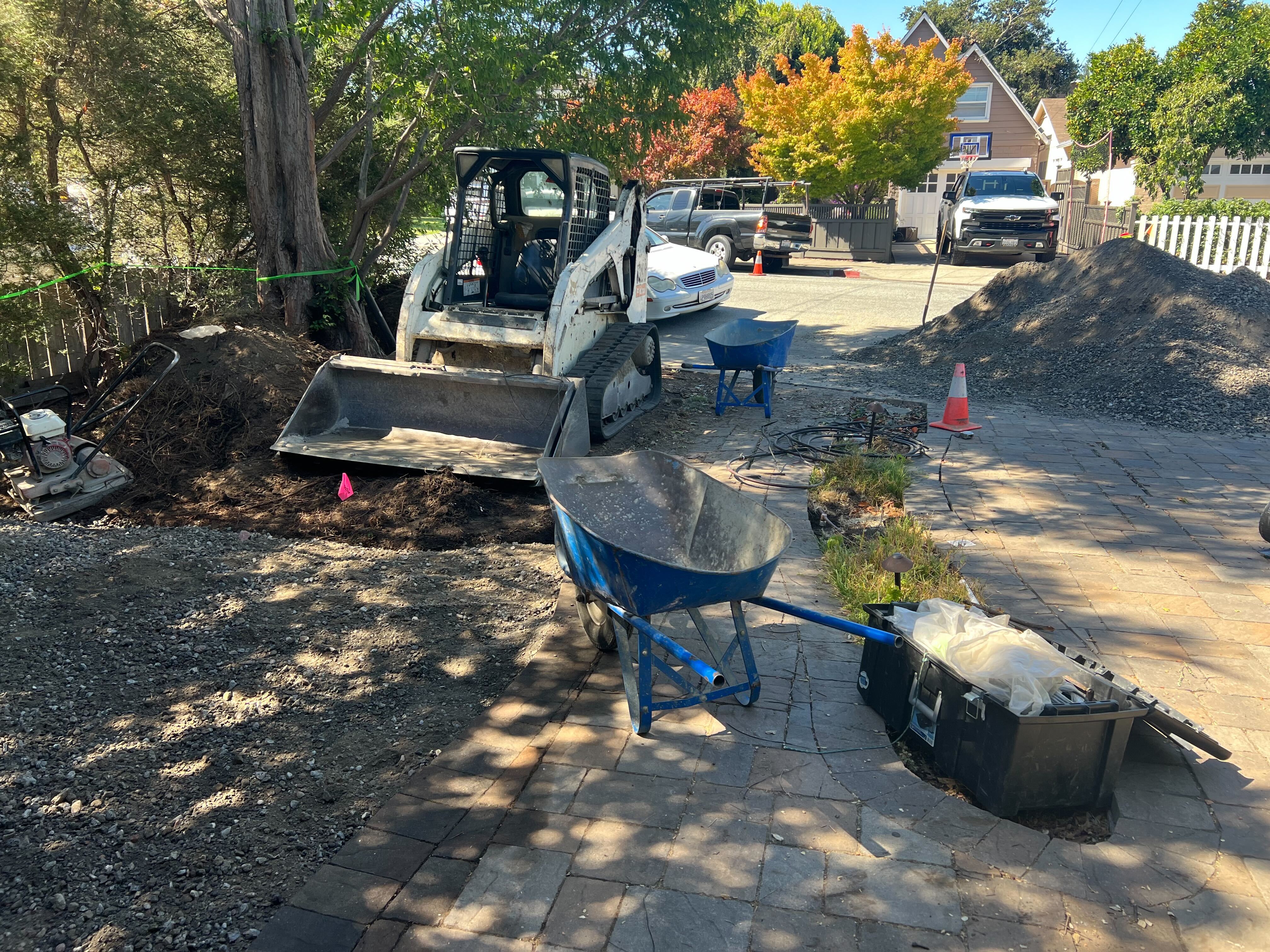 Site photo showing active base preparation for a paver driveway/approach with a skid steer, wheelbarrows and a large...