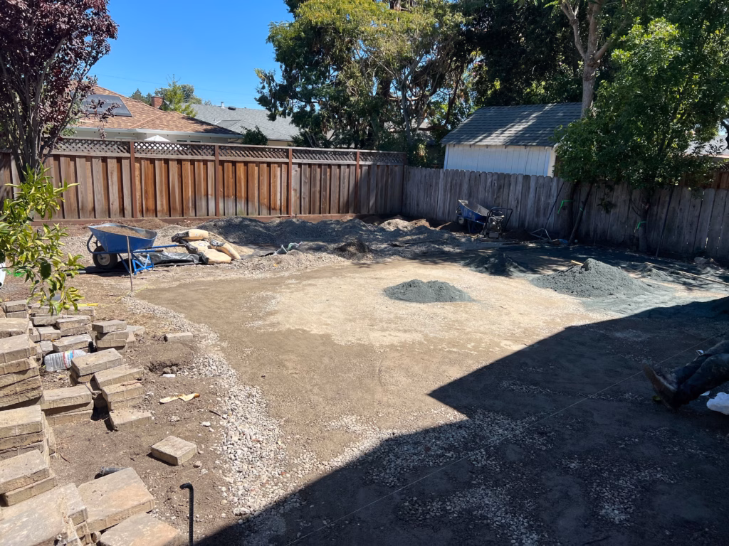 Backyard has been stripped to a compacted subgrade with piles of base material and bedding sand staged for a paver patio....