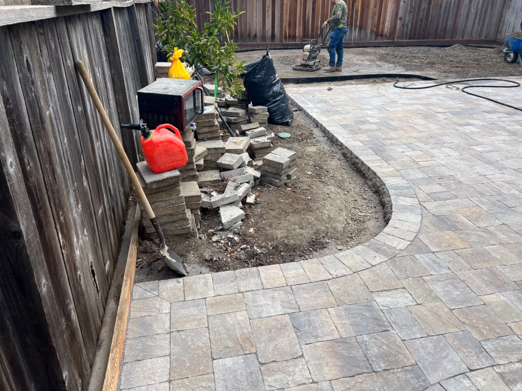 Partially installed interlocking paver patio in a backyard with a curved edge and exposed base. Stacked pavers, tools,...