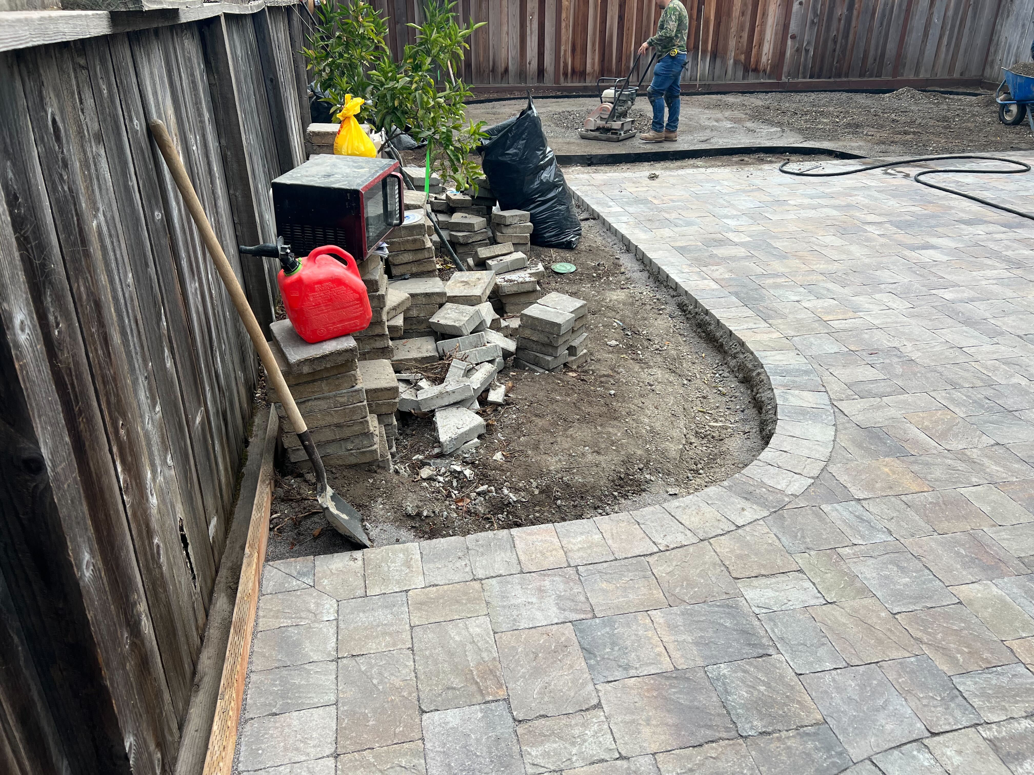 Partially installed interlocking paver patio in a backyard with a curved edge and exposed base. Stacked pavers, tools,...