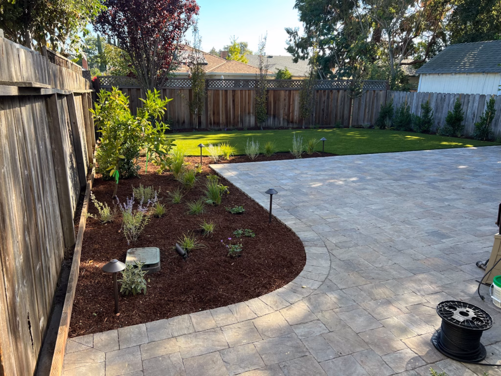 Backyard shows a newly installed modular concrete paver patio with a curved mulch planting bed and adjoining lawn....