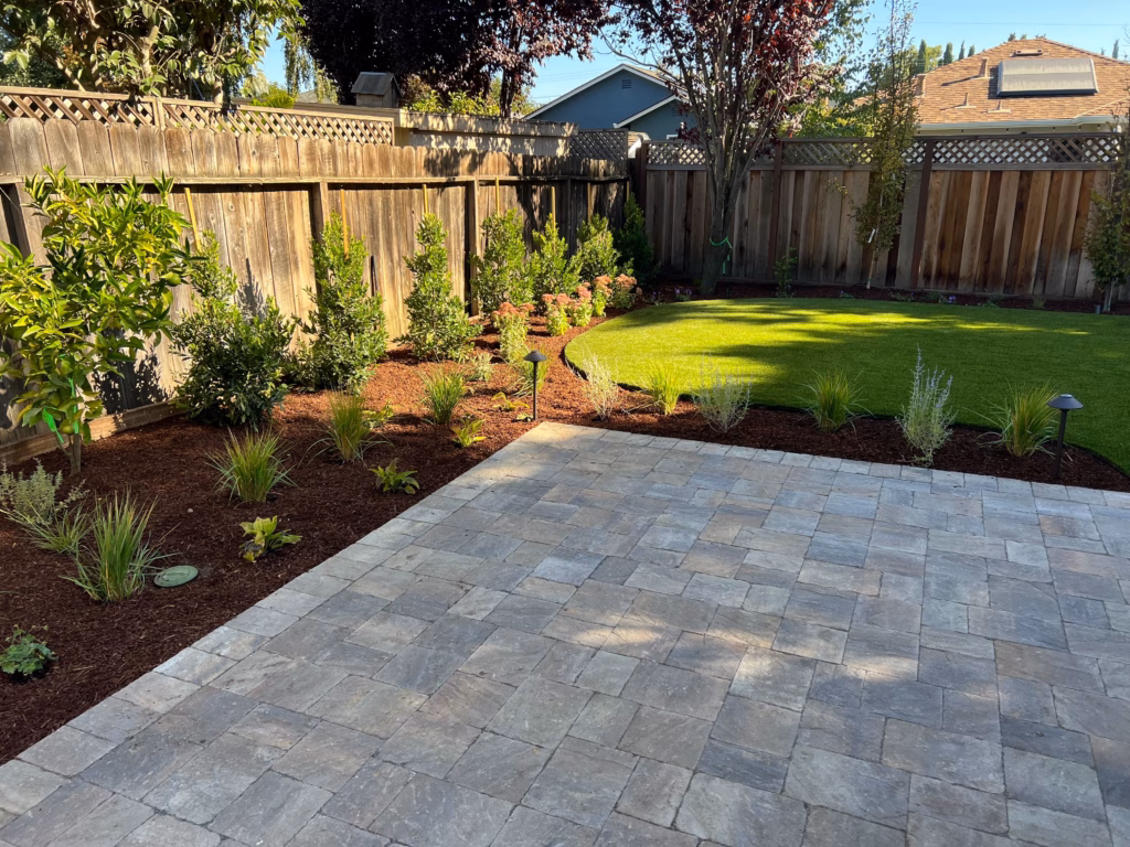 Finished backyard showing a newly installed interlocking paver patio transitioning to a fresh artificial turf lawn with...