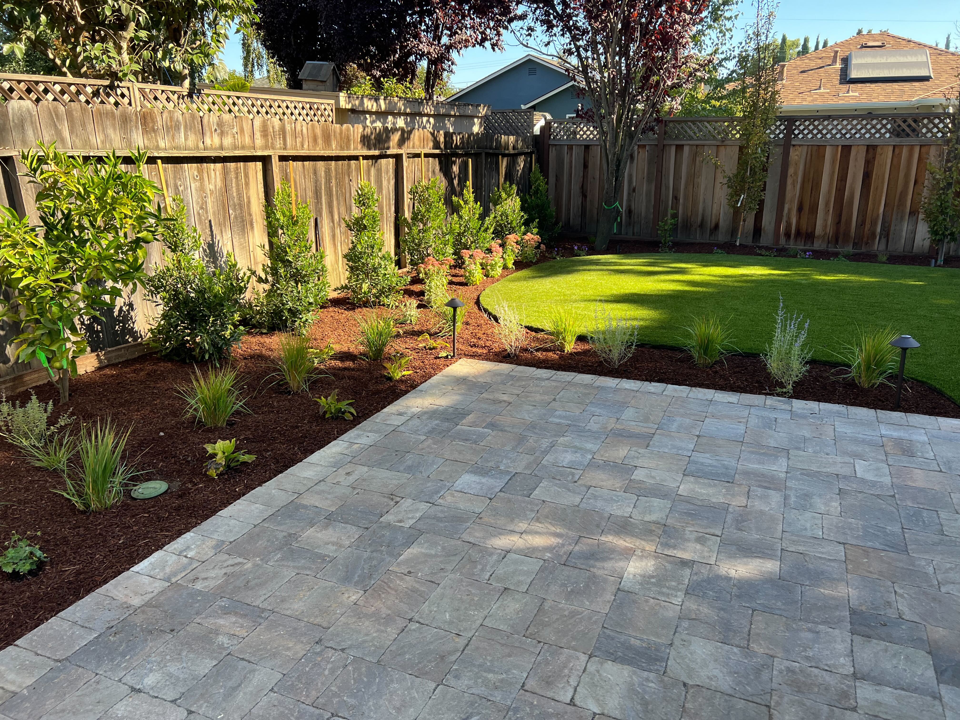 Finished backyard showing a newly installed interlocking paver patio transitioning to a fresh artificial turf lawn with...