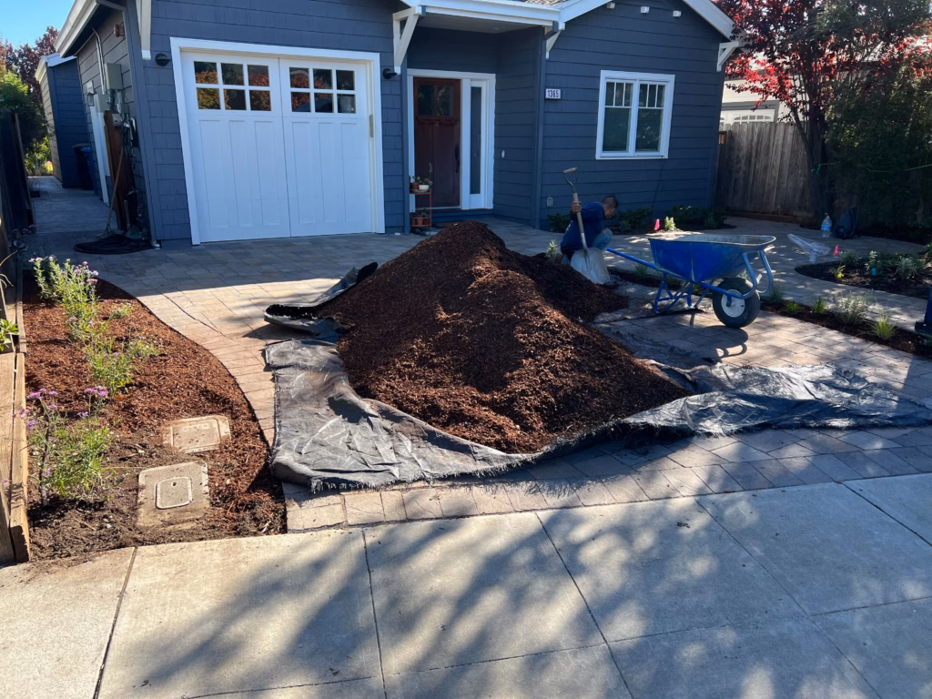 Front yard shows a newly installed interlocking paver driveway/patio area with a large pile of mulch/soil on a tarp and a...