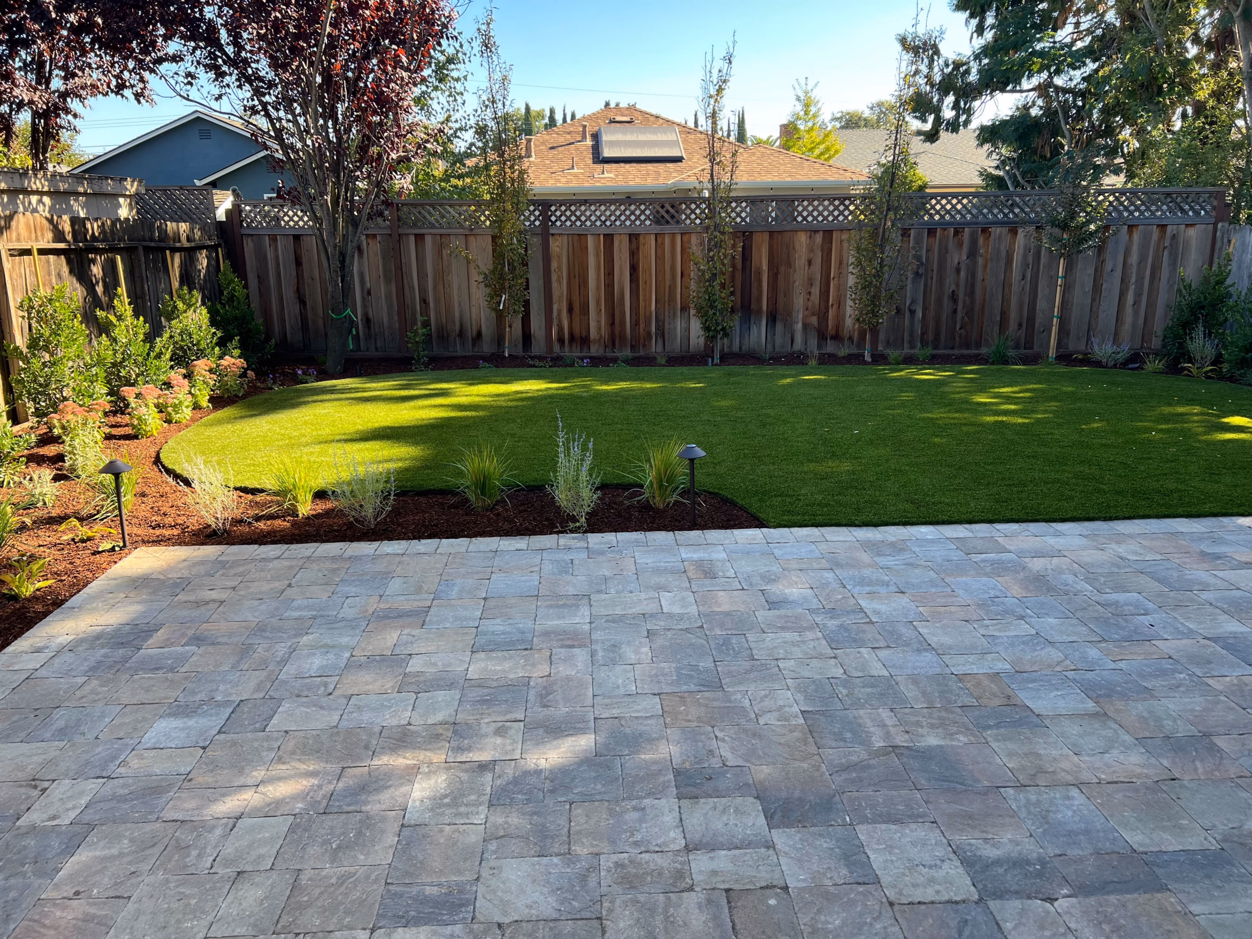 Finished backyard showing a newly installed interlocking paver patio in the foreground and an artificial turf lawn with...