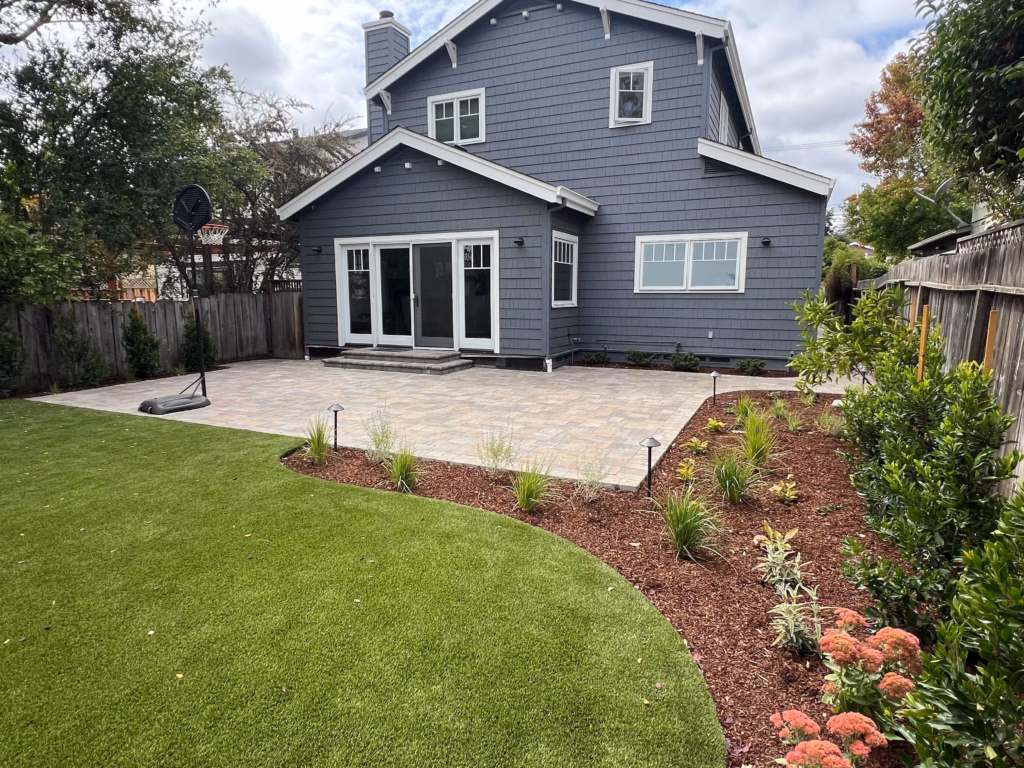 Finished interlocking paver patio installed against the rear of a two-story residence, showing clean joints and a...