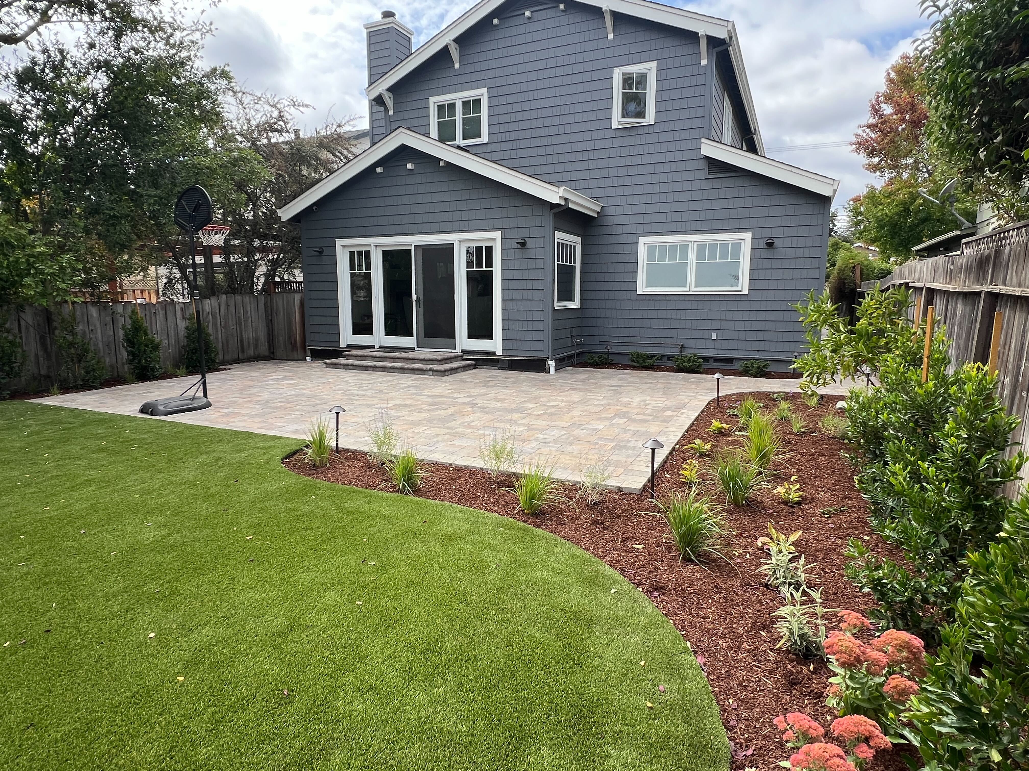 Finished interlocking paver patio installed against the rear of a two-story residence, showing clean joints and a...