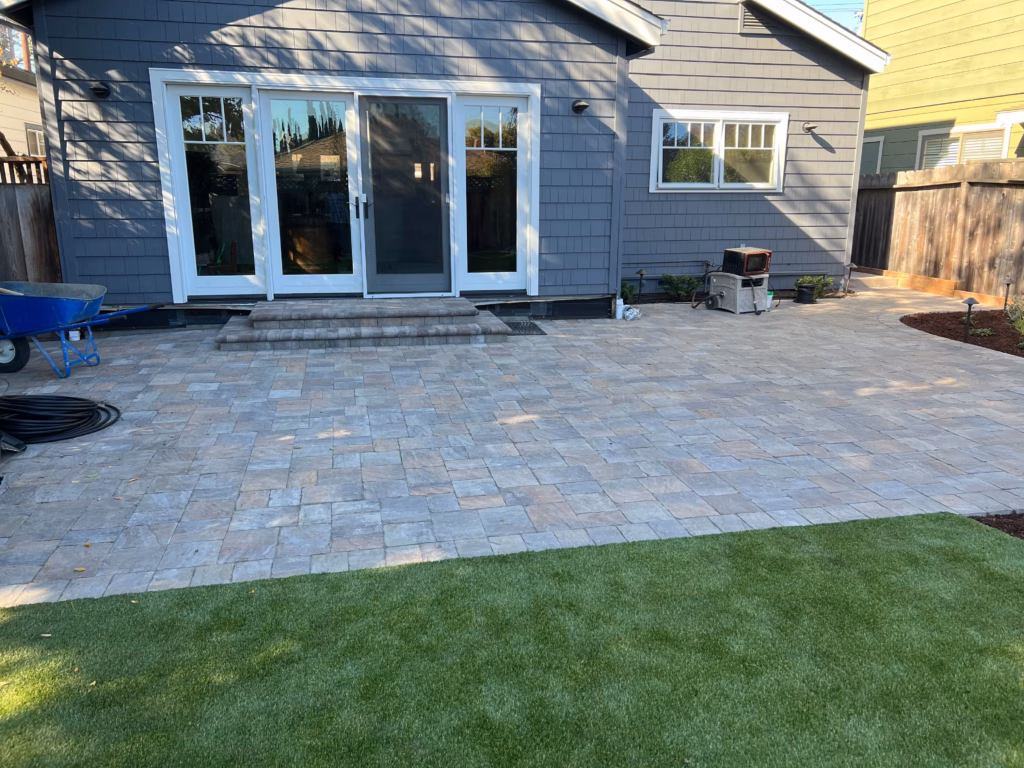Rear residential yard showing a completed interlocking concrete paver patio abutting a strip of artificial turf. Paver...