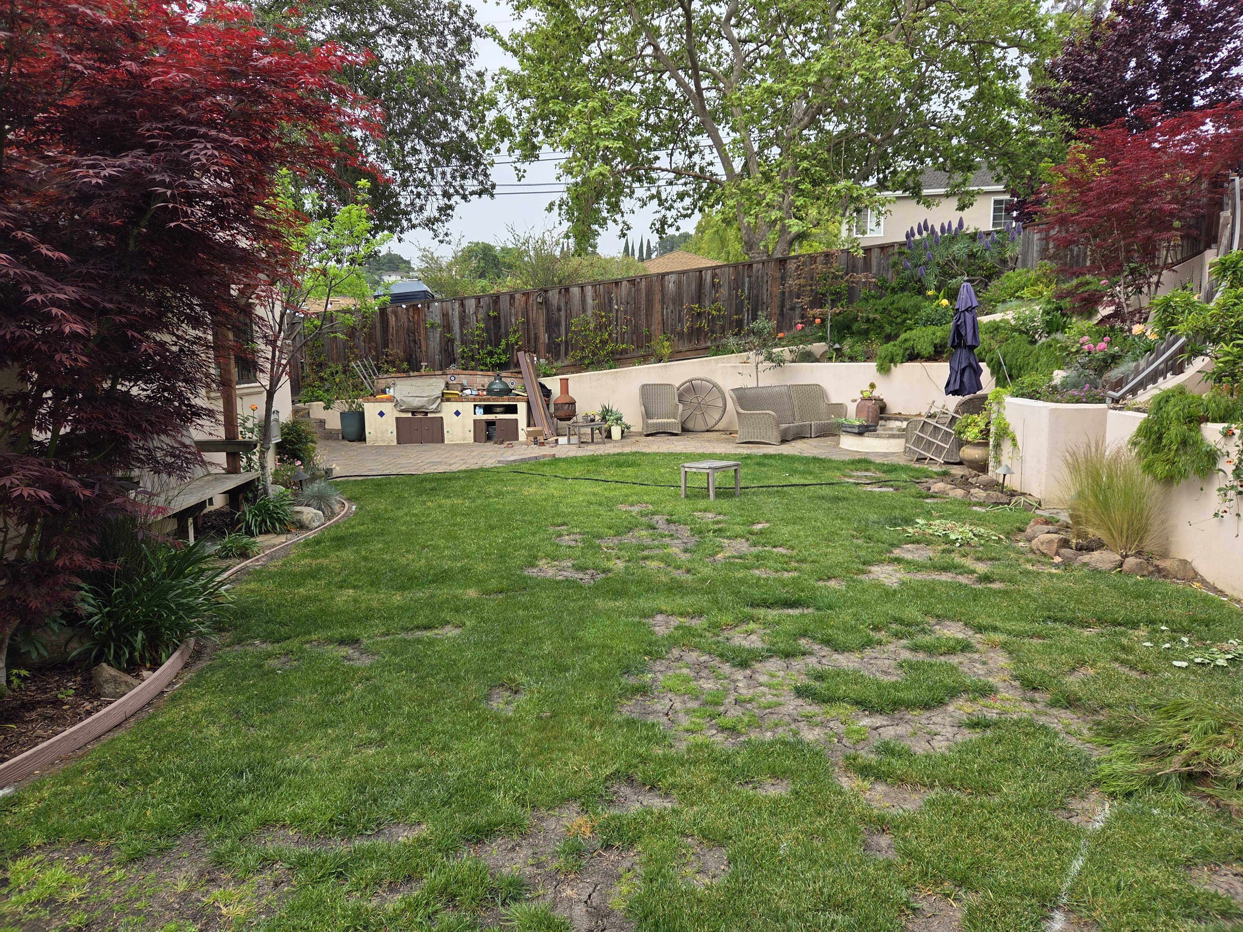 Residential backyard showing a patchy natural lawn with worn areas in front of a tiered segmental retaining wall. The...