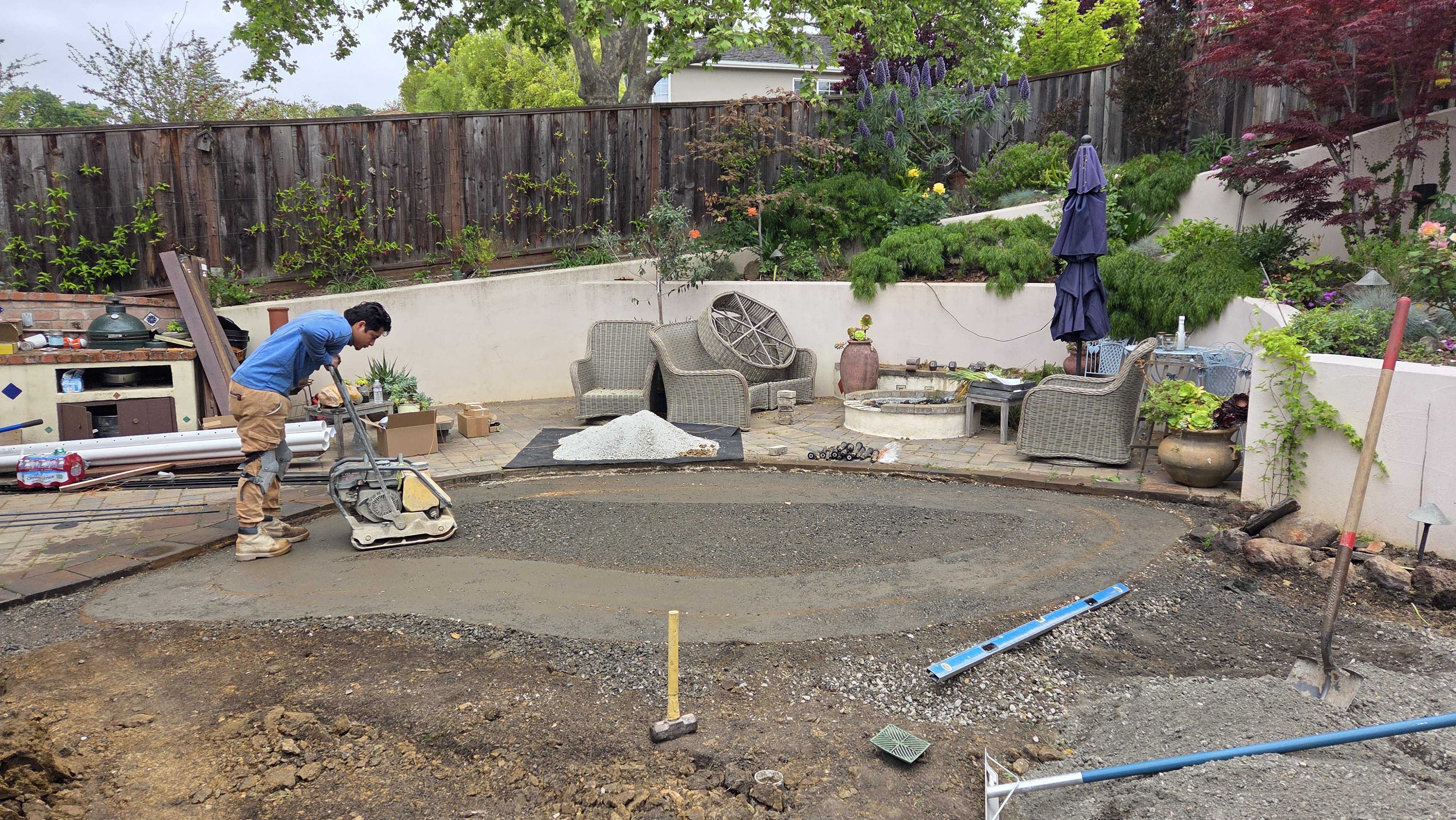 Worker operating a plate compactor to finish an aggregate base for a future paver patio in a residential backyard....