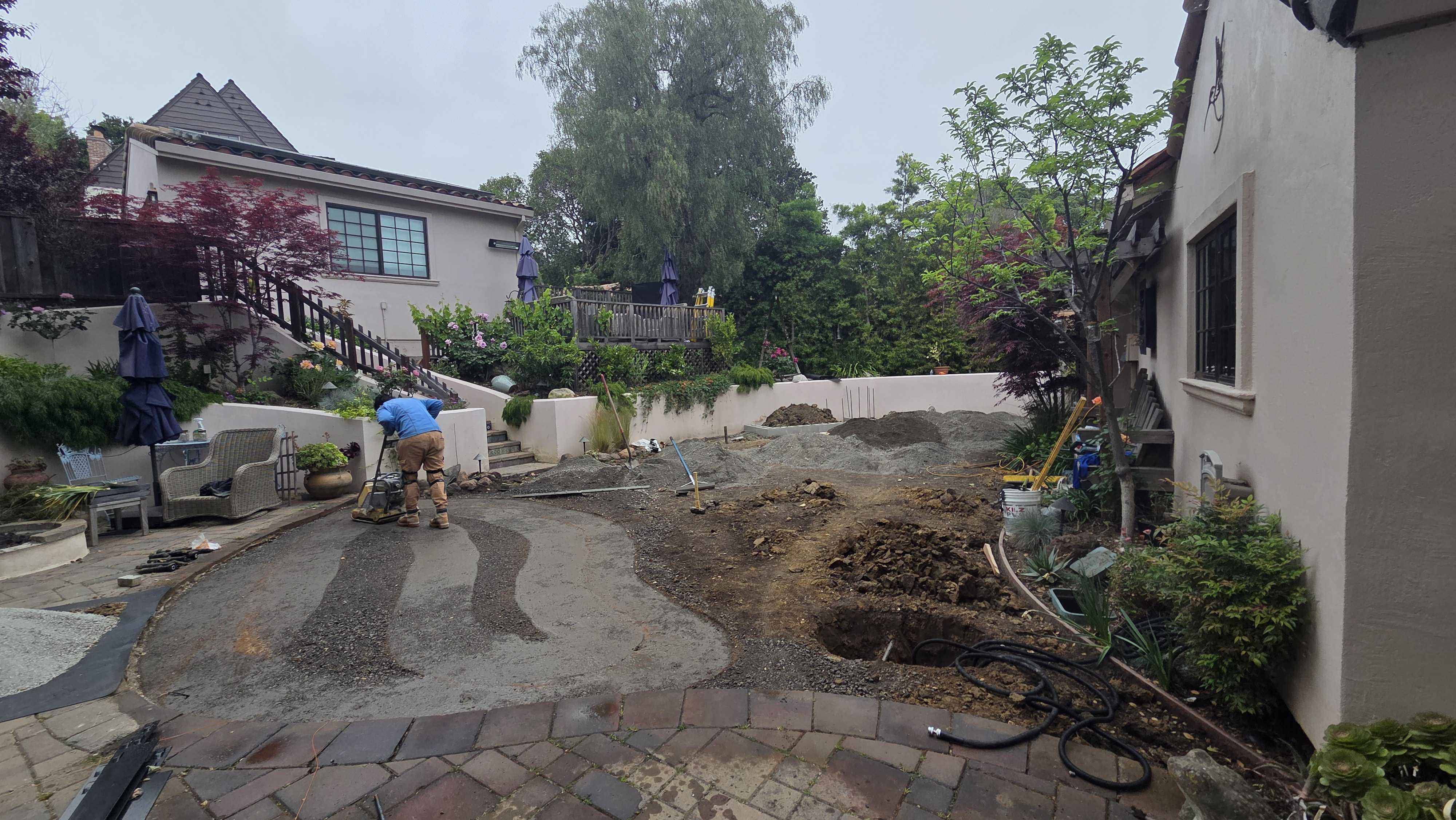 Backyard patio area with existing pavers removed and base preparation underway; crew member is grading/compacting the...