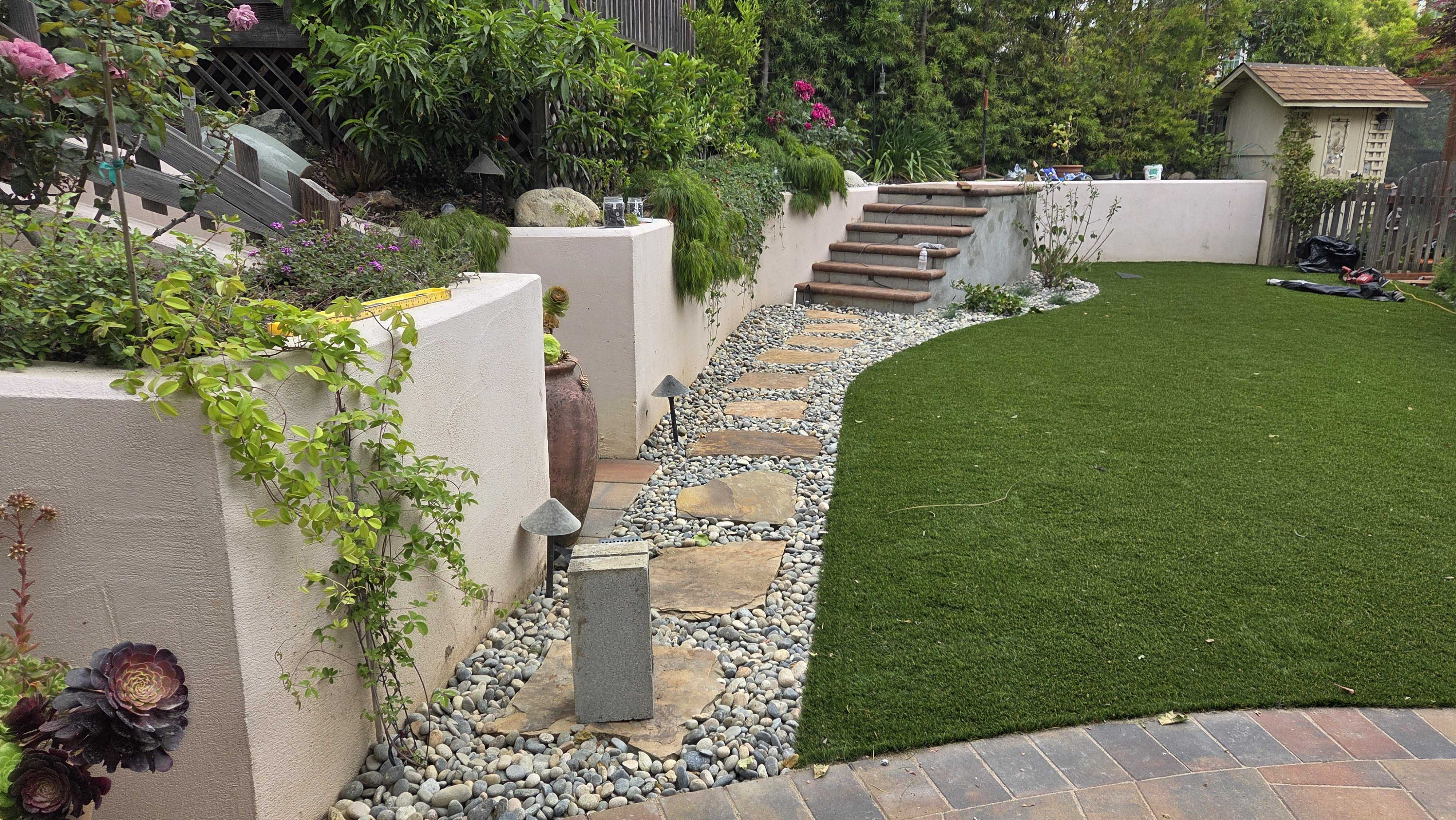 Completed backyard installation showing an interlocking paver patio edging next to a curved artificial turf lawn. A...