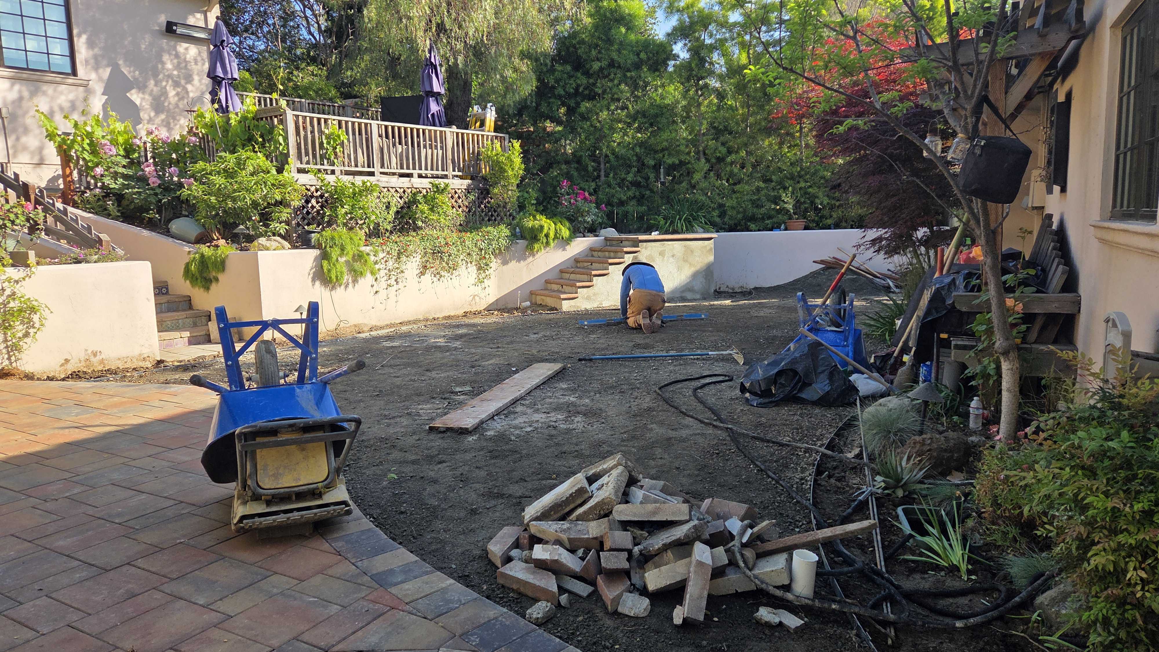 Backyard renovation scene showing an existing paver patio edge and a cleared area with exposed subgrade where crew are...