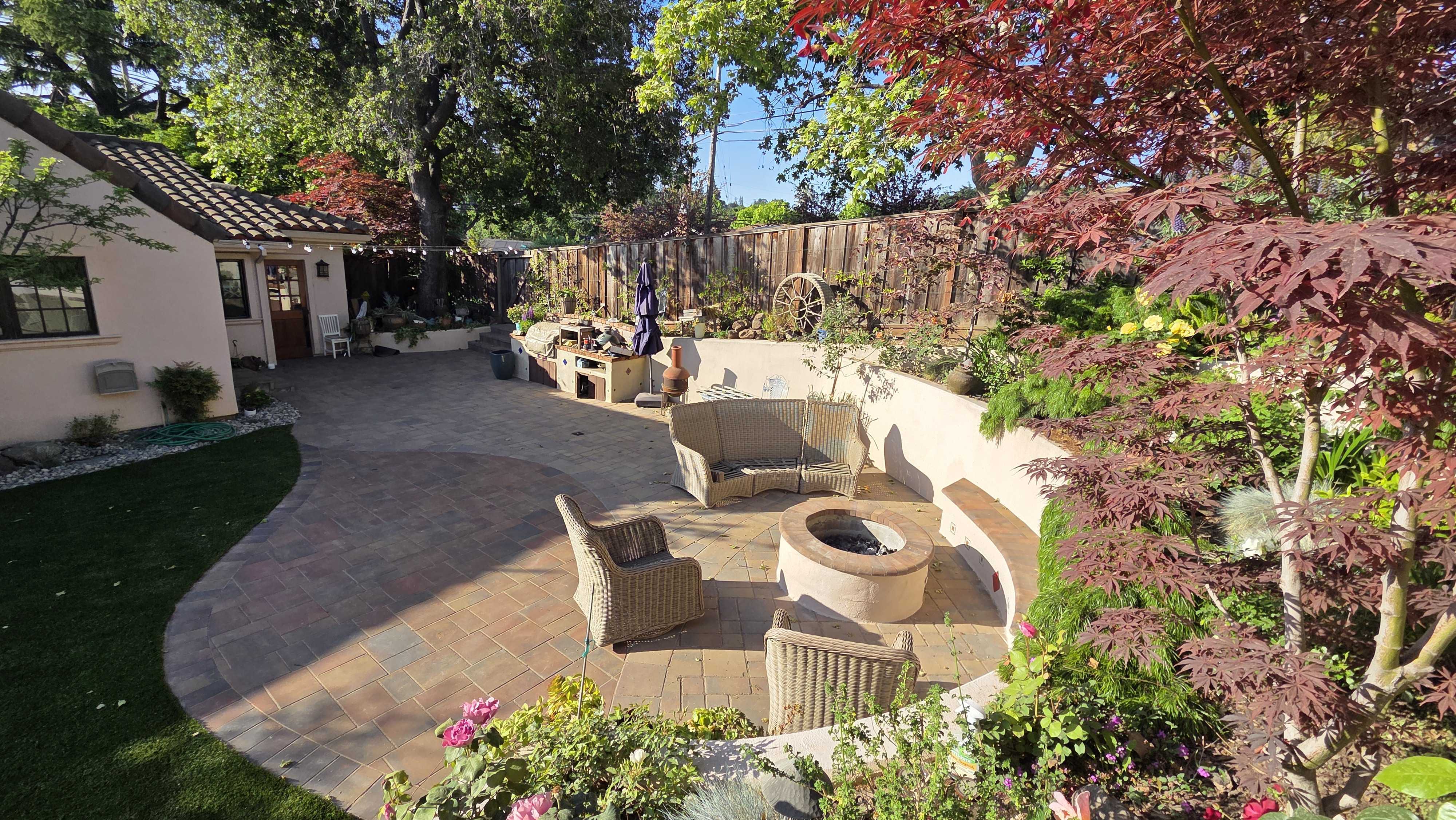 Finished residential backyard showing an interlocking paver patio with a central circular fire pit and curved segmental...