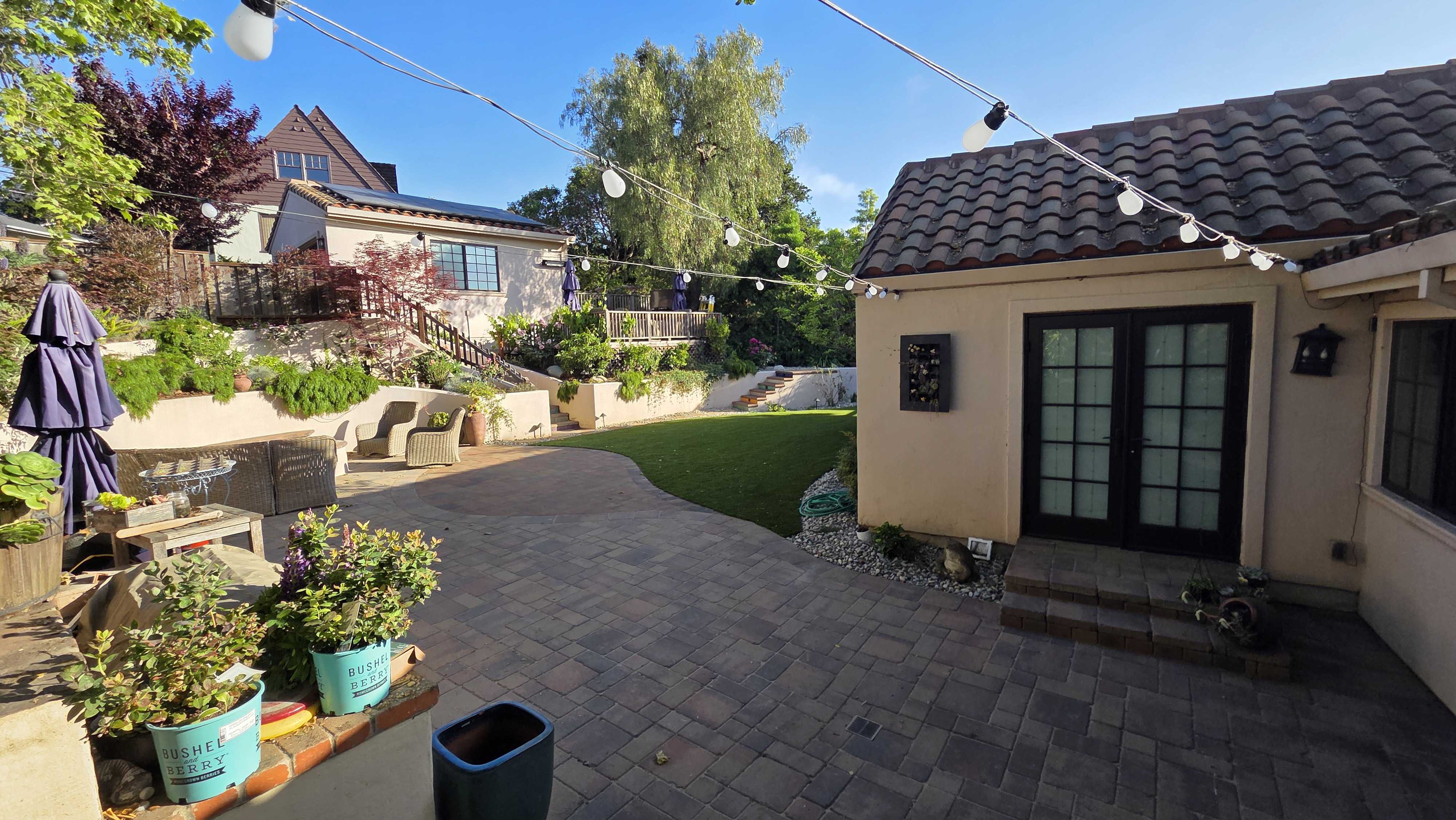 Finished backyard showing a large interlocking paver patio flowing into a curved artificial turf lawn. Segmental...