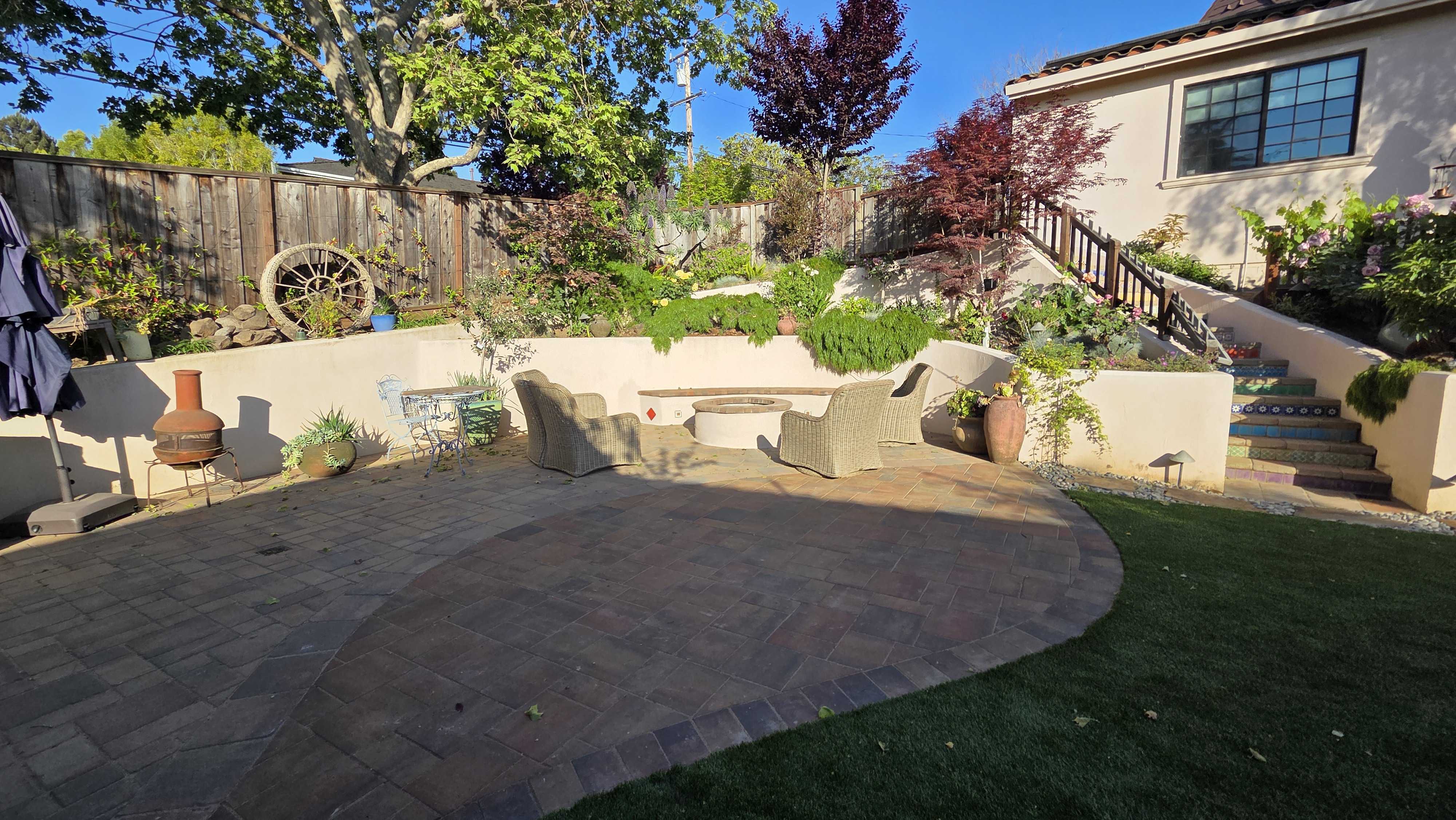 Finished backyard showing a curved interlocking paver patio adjacent to a strip of artificial turf. A low segmental...