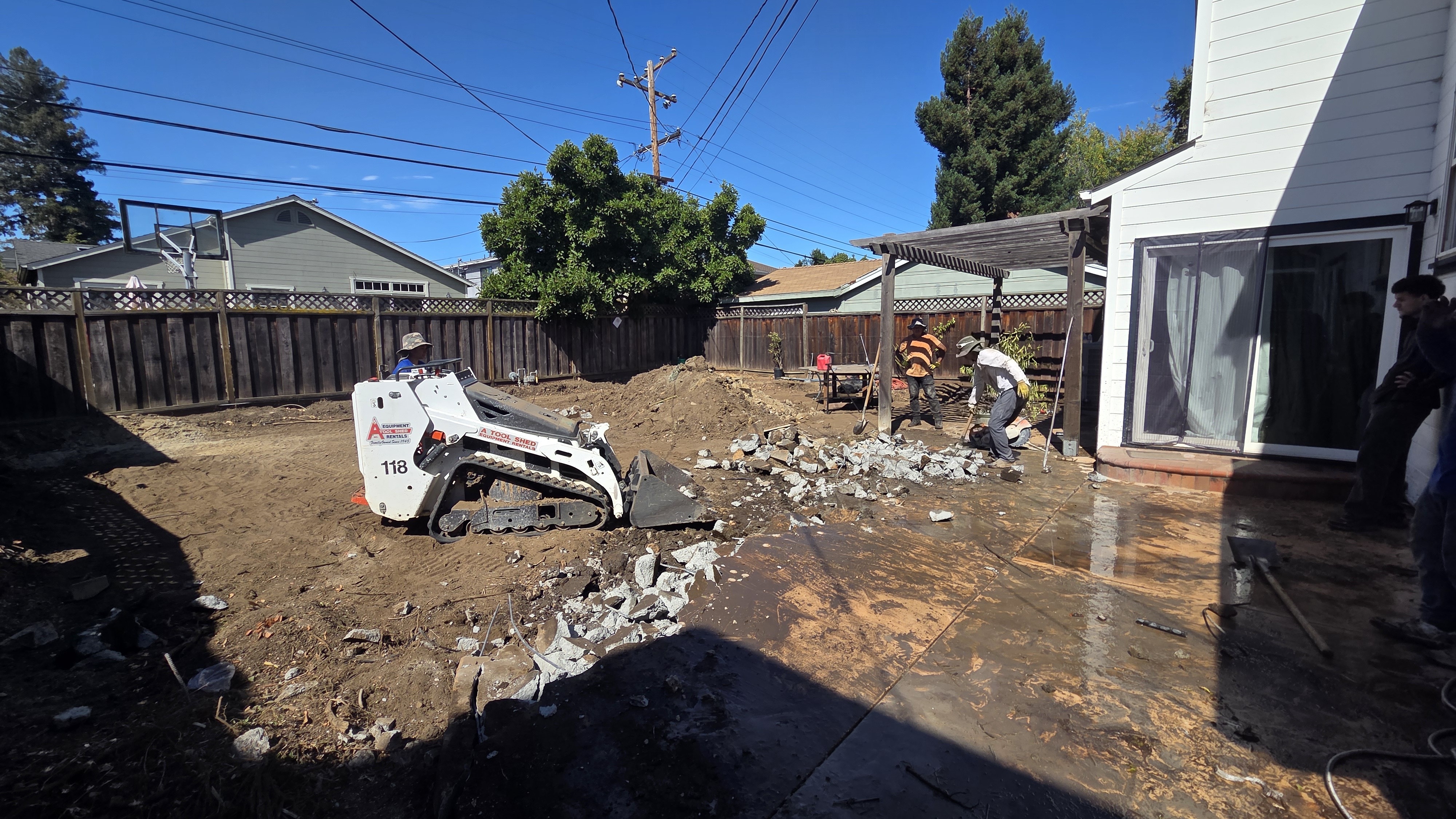 Backyard concrete patio is actively being demolished; a mini skid steer is moving broken slab while crew members break up...