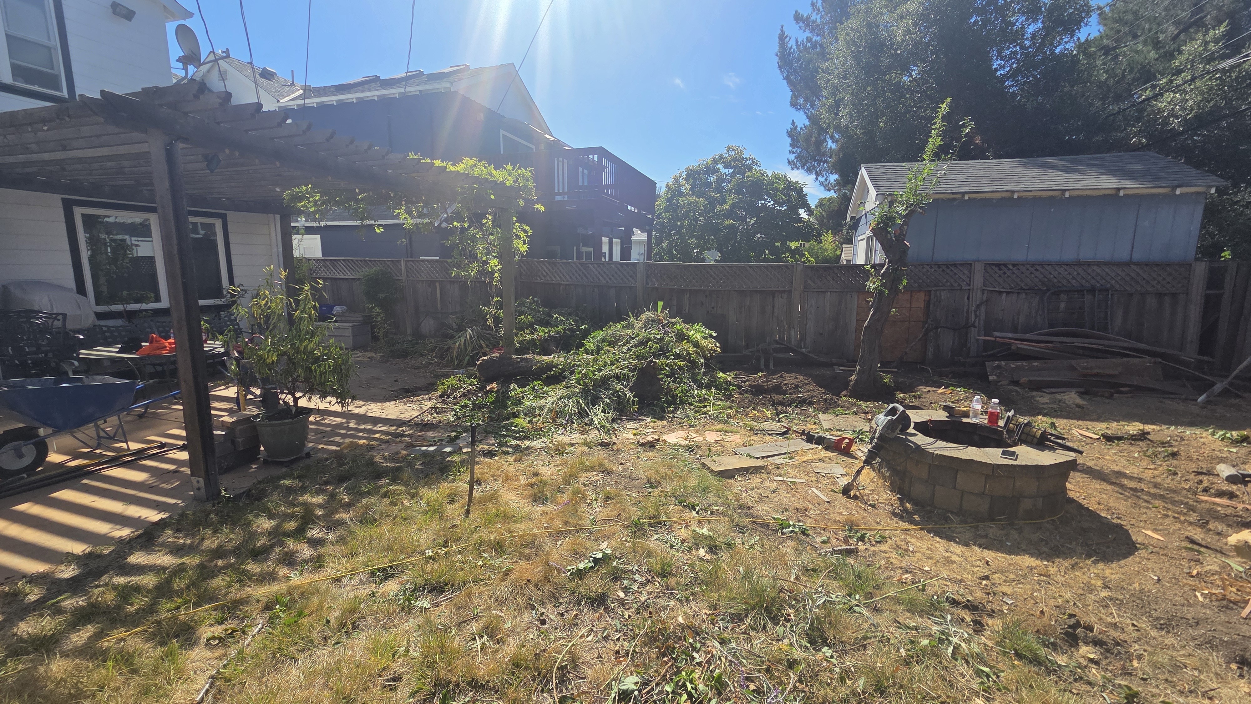 Backyard site photo showing a patchy natural lawn with bare dirt and a circular block fire pit in the yard center....