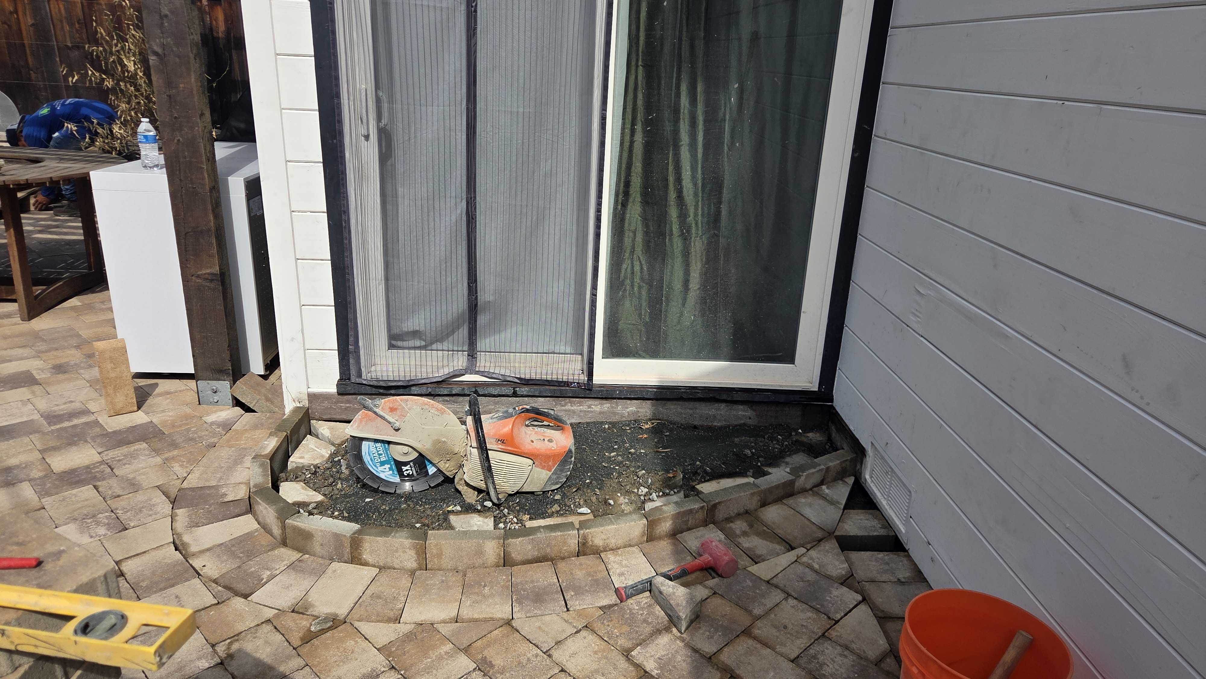 Worker-stage photo of an interlocking paver patio being installed at a sliding glass door. A curved segmental knee...