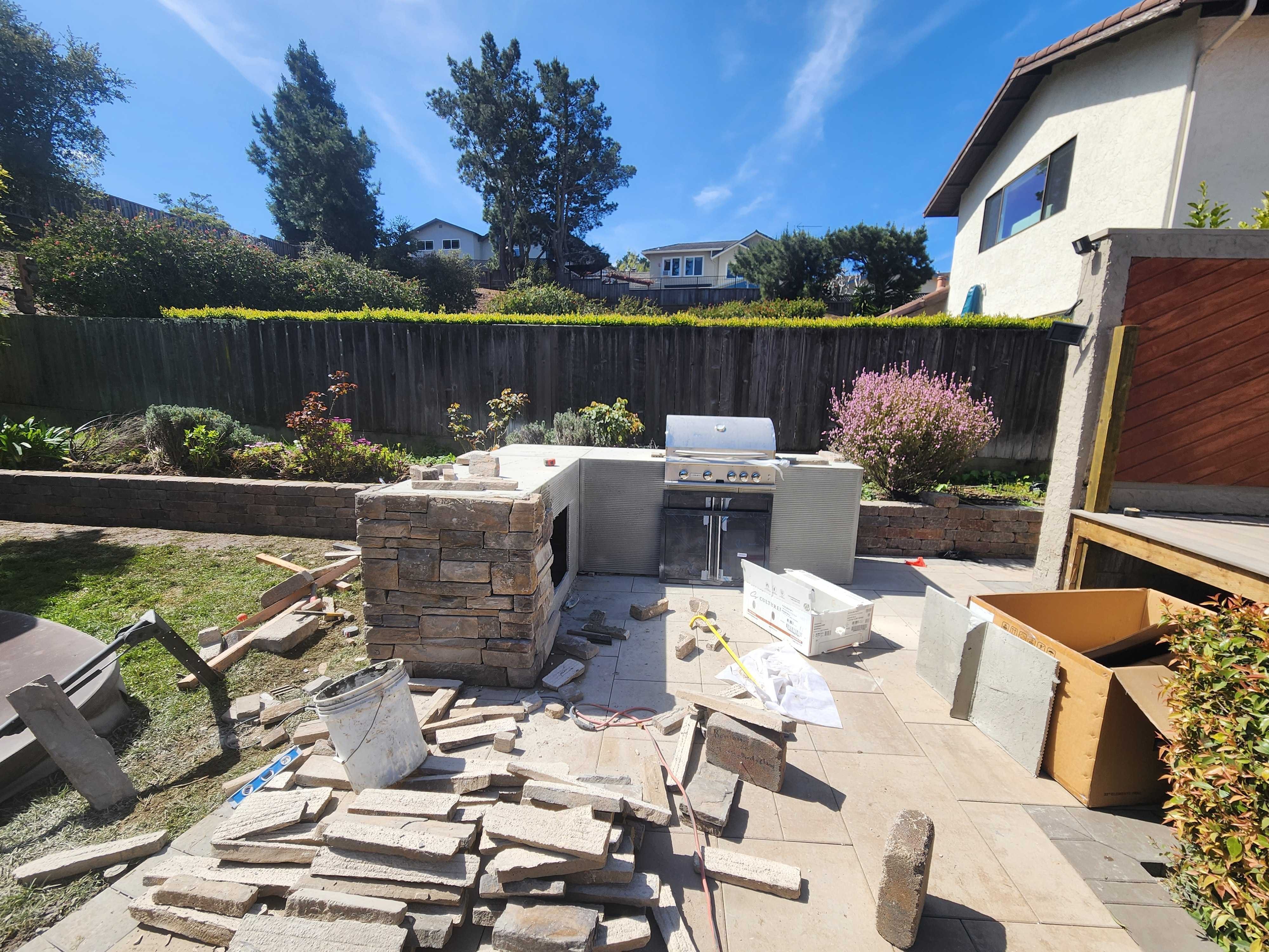 Outdoor kitchen island mid-build: one wing wrapped in stacked-stone veneer, the stainless grill already set in the cement-board run.