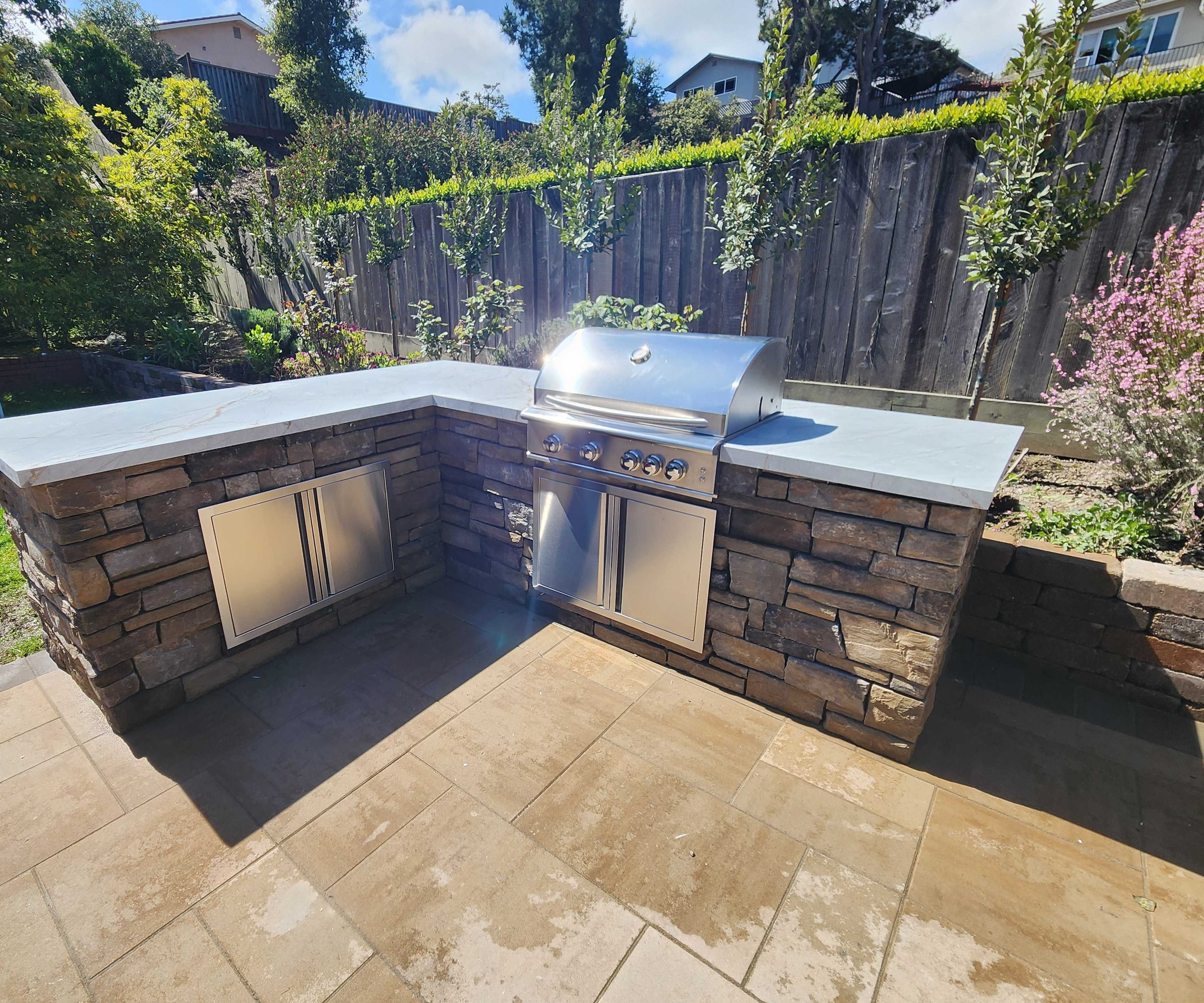 L-shape stacked-stone outdoor kitchen with a stainless grill and honed gray countertop on the new paver patio.