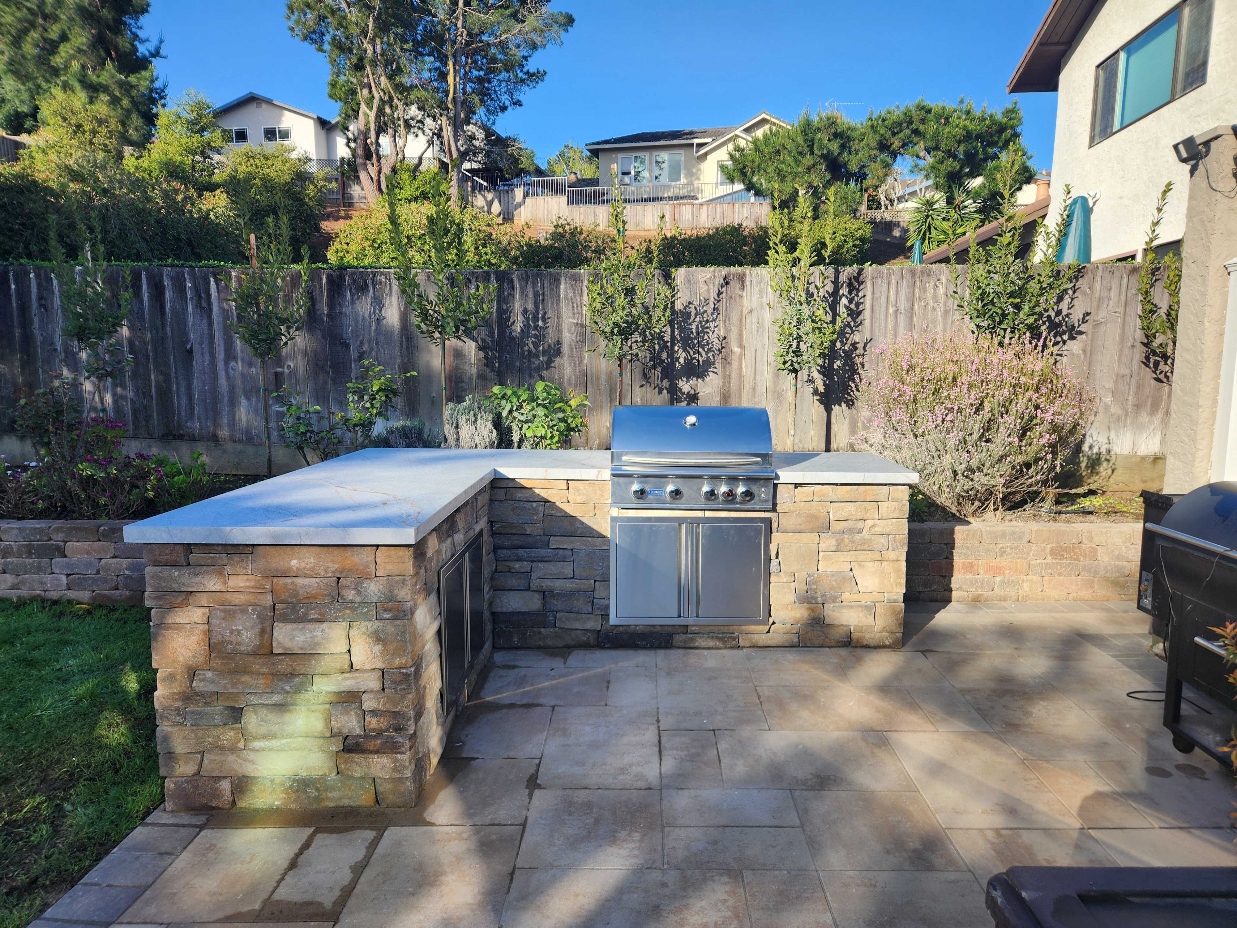 Stacked-stone outdoor kitchen with stainless grill and honed stone counter in warm evening light, on the paver patio