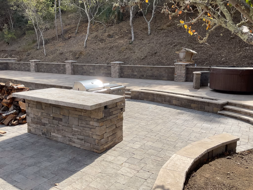 Wide backyard view with paver patio, stone-veneer retaining wall, capped pillars, outdoor kitchen island and hot tub...