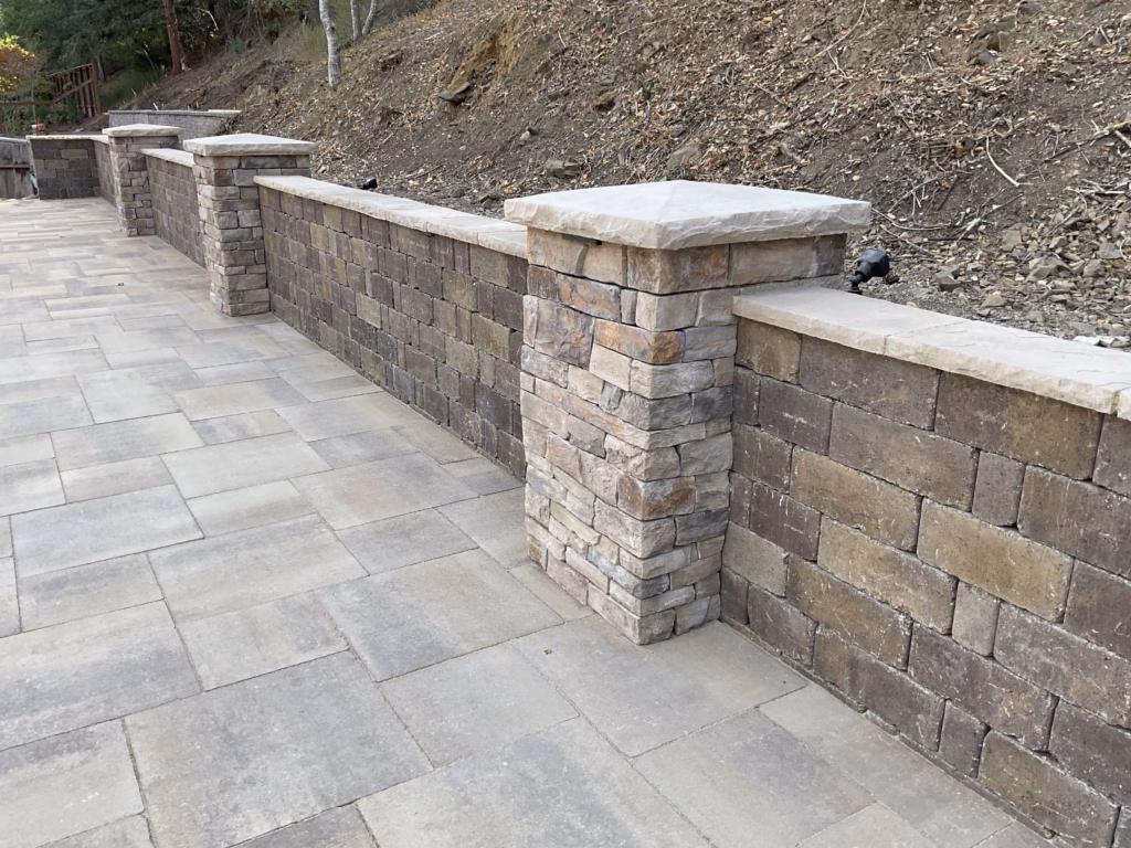Stone-veneer segmental retaining wall with capped pillars along the hillside edge of a paver patio