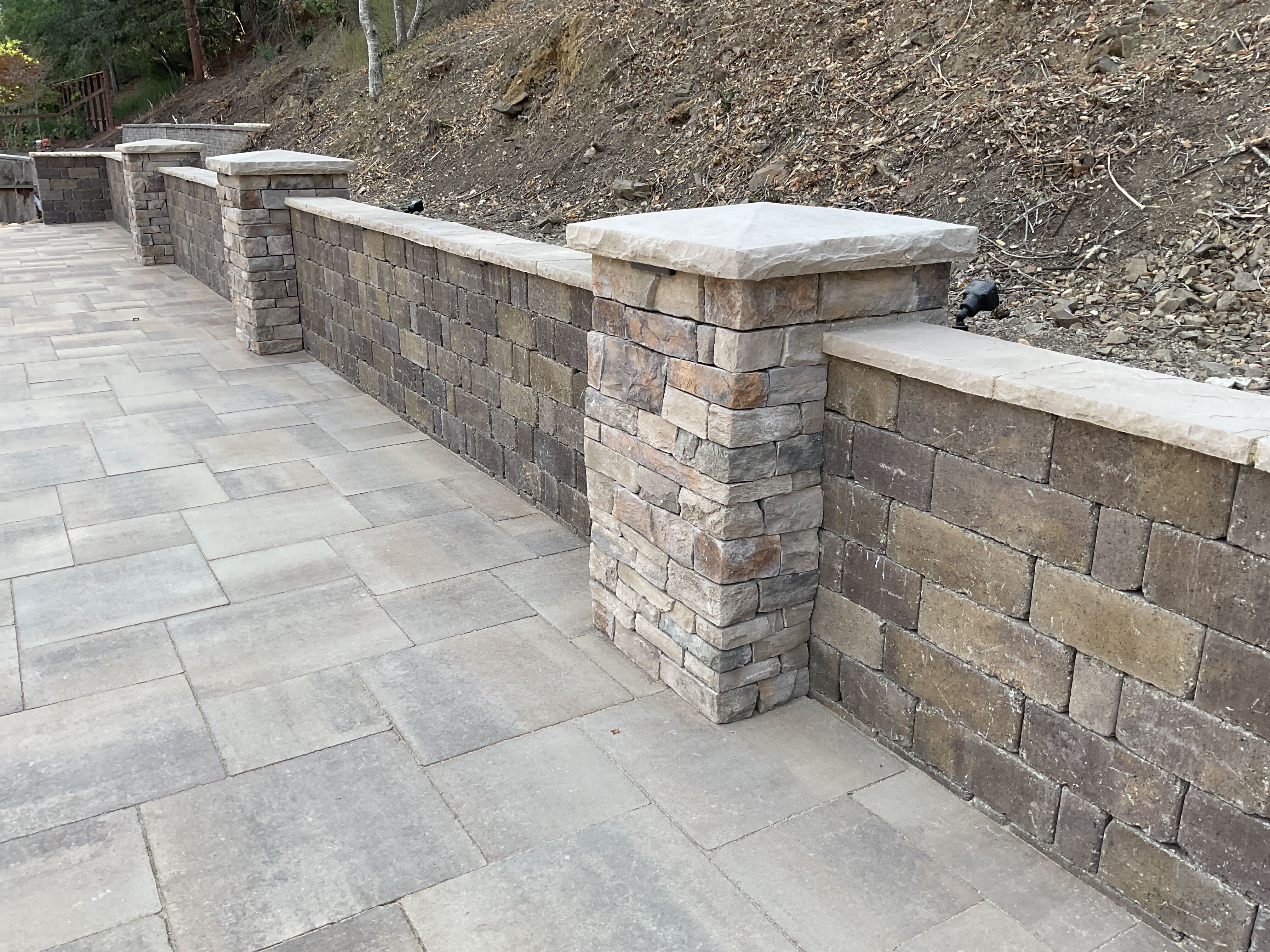 Stone-veneer segmental retaining wall with capped pillars along the hillside edge of a paver patio