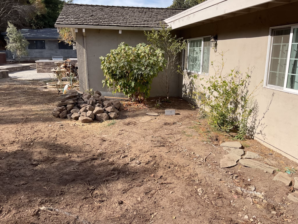 Before — Backyard before construction: cleared bare-dirt area with rock pile, mature shrubs along the rear of the home and the...