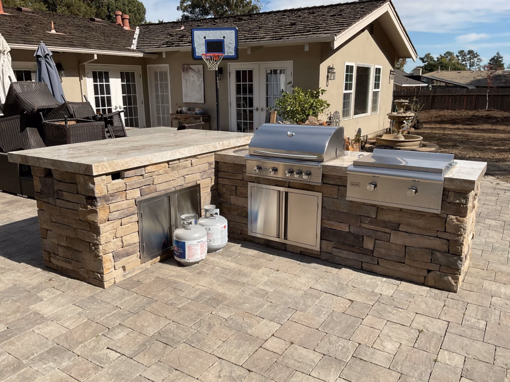 Stone-veneer outdoor kitchen island with built-in stainless steel grill on a paver patio with the home and hillside in...