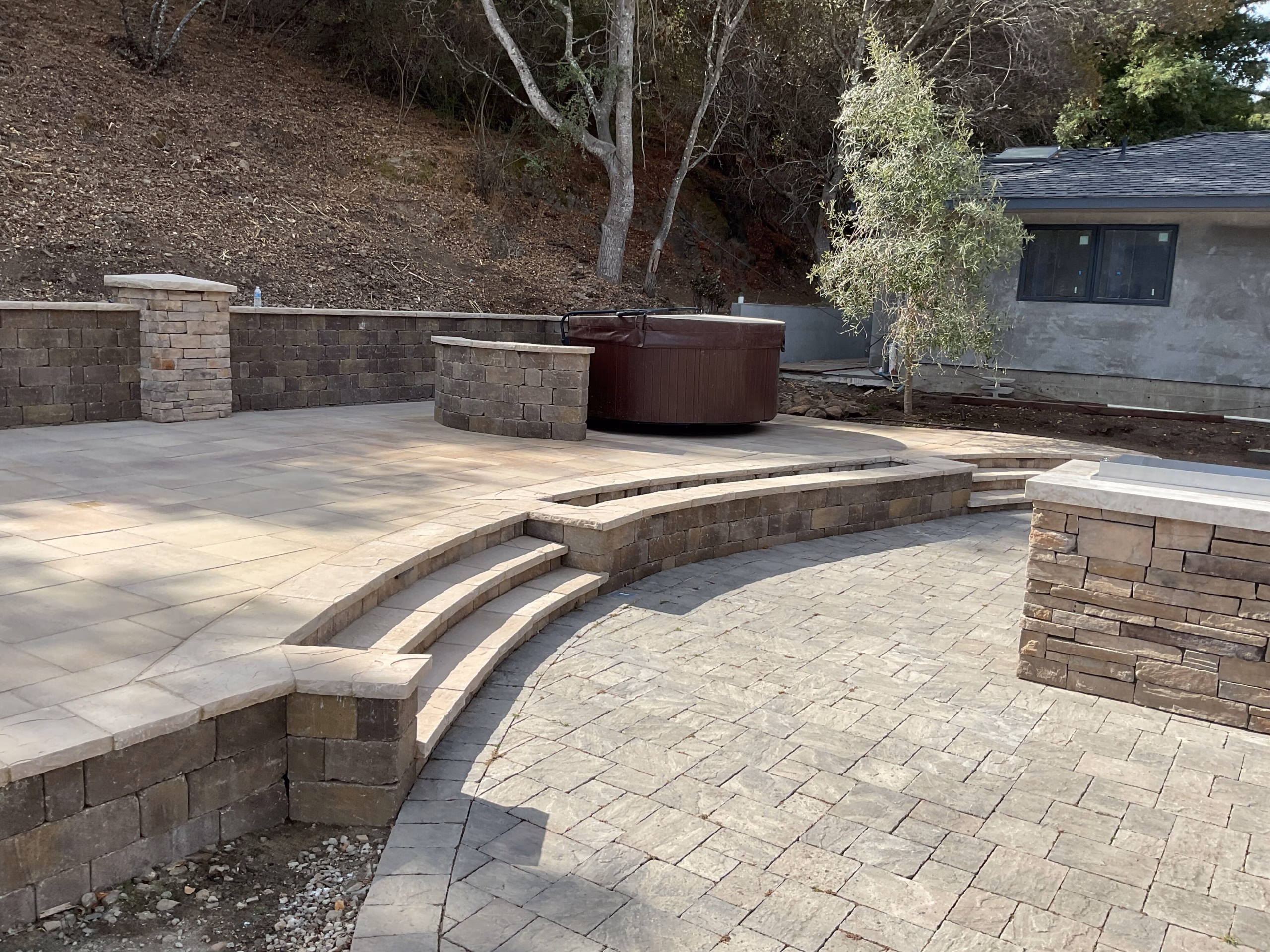 Tiered paver patio with curved bullnose steps, a segmental retaining wall and a capped pillar on a San Mateo hillside.
