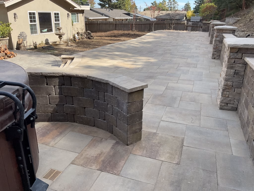 Wide paver patio with curved seat wall, segmental retaining wall and capped pillars, hot tub at left edge