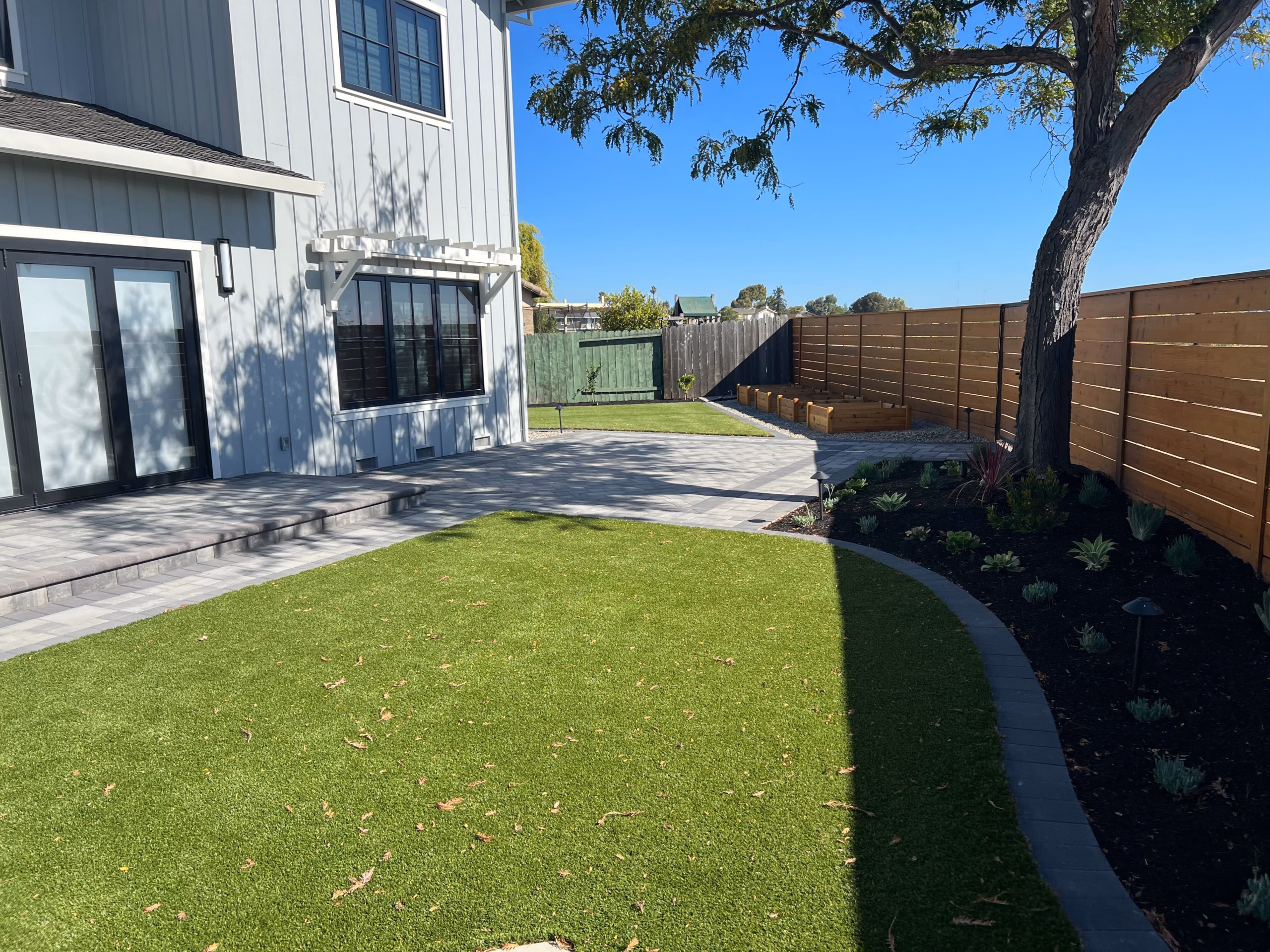 An artificial-turf lawn edged by a charcoal paver border, a planted strip along a new horizontal-board fence and a paver patio at the house, in Redwood City
